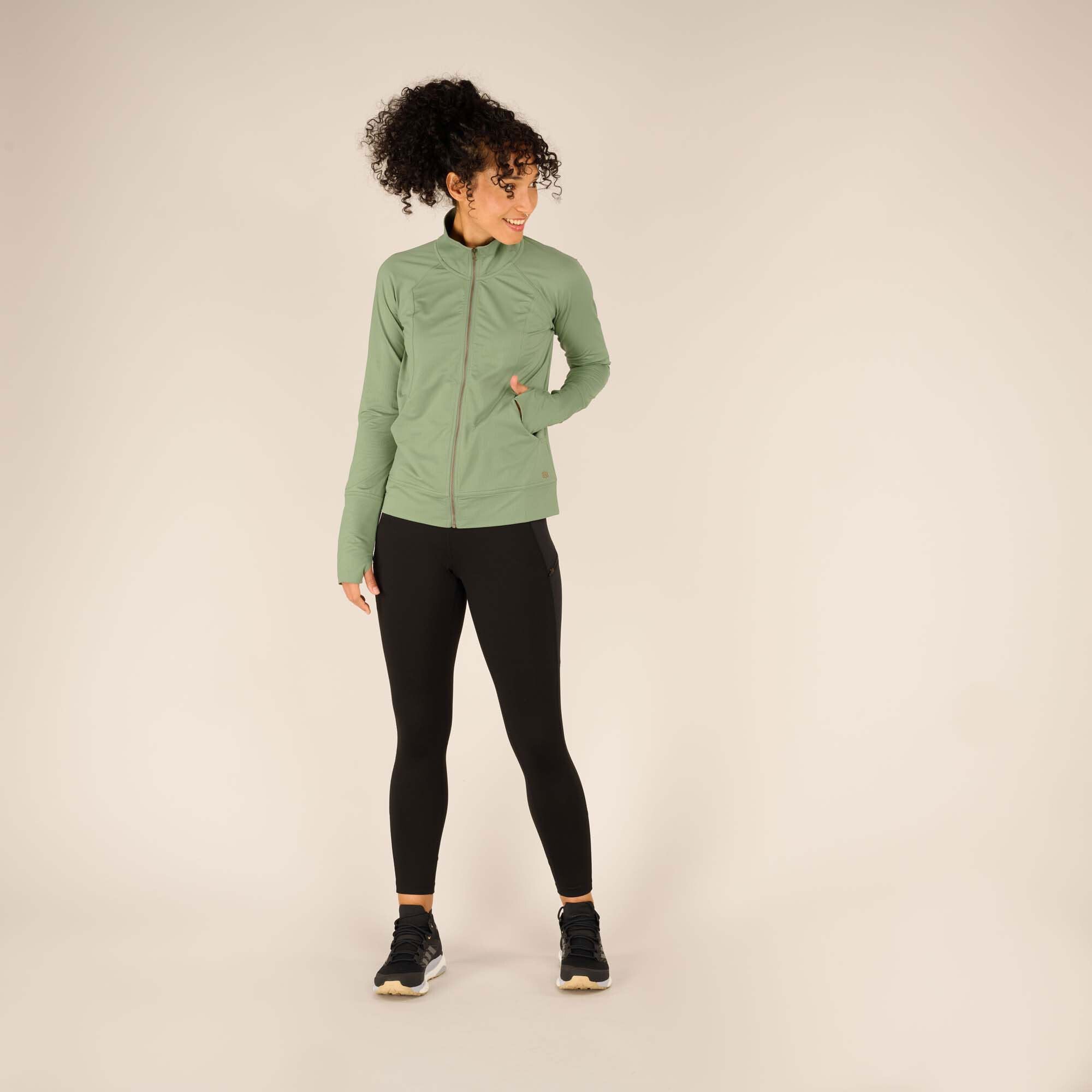 A full-body shot of a model wearing the Sherpa Adventure Gear Aarti Full Zip Jacket in Green. The model stands in a neutral pose with one hand in her pocket, smiling slightly to the side. She pairs the jacket with black leggings and black sneakers. The background is a clean, beige studio setting.