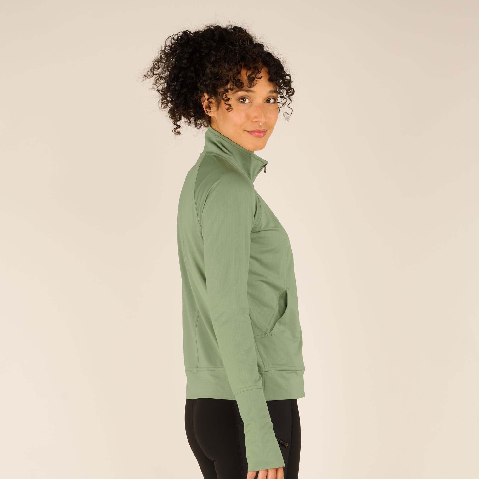 A side view of the model wearing the Sherpa Adventure Gear Aarti Full Zip Jacket in Green, showing the jacket's slim fit and structured design. The model’s curly hair is tied back, and the jacket’s collar is zipped up to the top. The side seam pocket is visible, and the sleeve features a thumbhole detail.