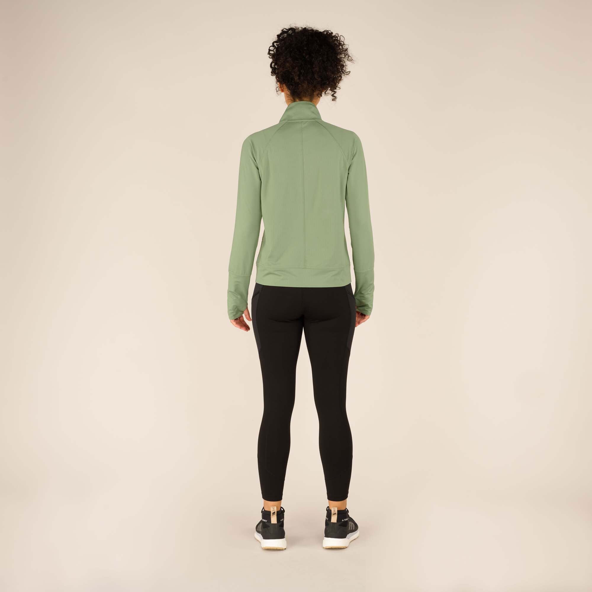 A rear view of the model wearing the Sherpa Adventure Gear Aarti Full Zip Jacket in Green, showcasing the simple back panel design and the fitted silhouette. The jacket extends slightly past the waist, and the sleeves fit snugly around the arms. The background is the same clean, beige studio setting.