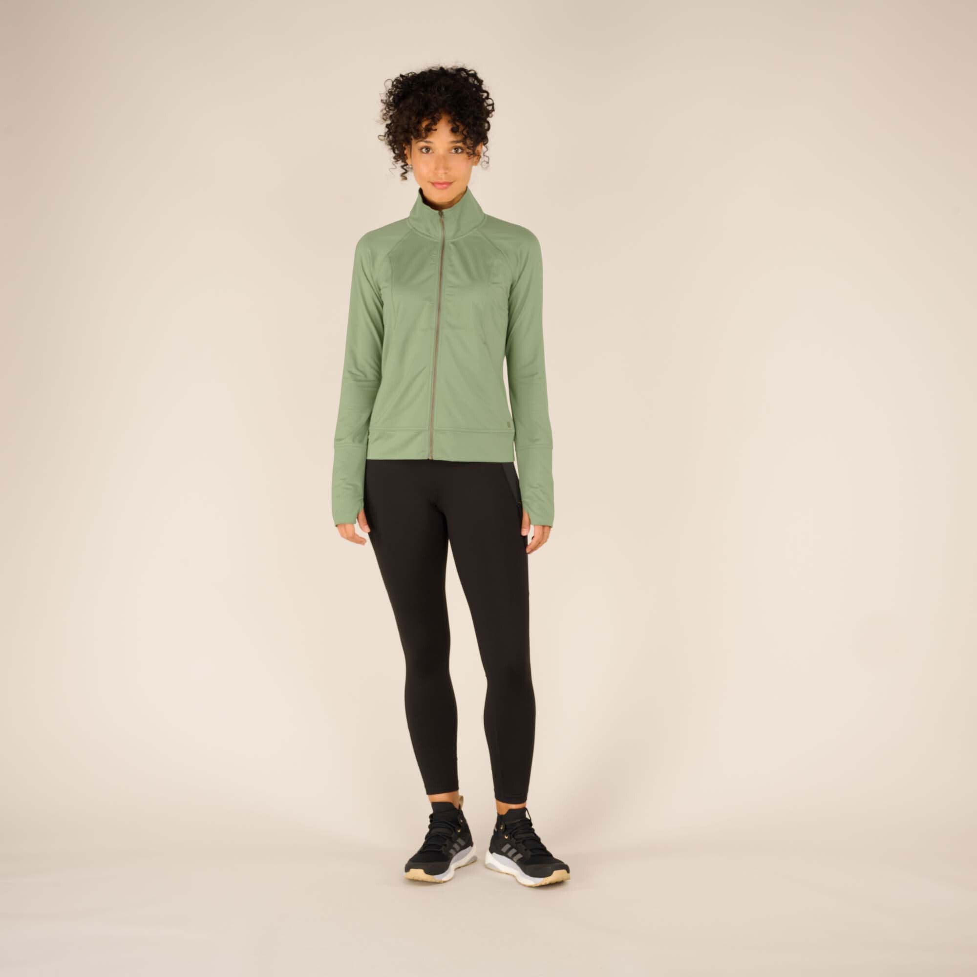 A front-facing image of the model standing with her arms relaxed by her sides, fully zipped in the Sherpa Adventure Gear Aarti Full Zip Jacket in Green. The jacket’s fitted cut and smooth, matte fabric texture are visible. She pairs the jacket with black leggings and black sneakers. The model maintains a neutral expression.