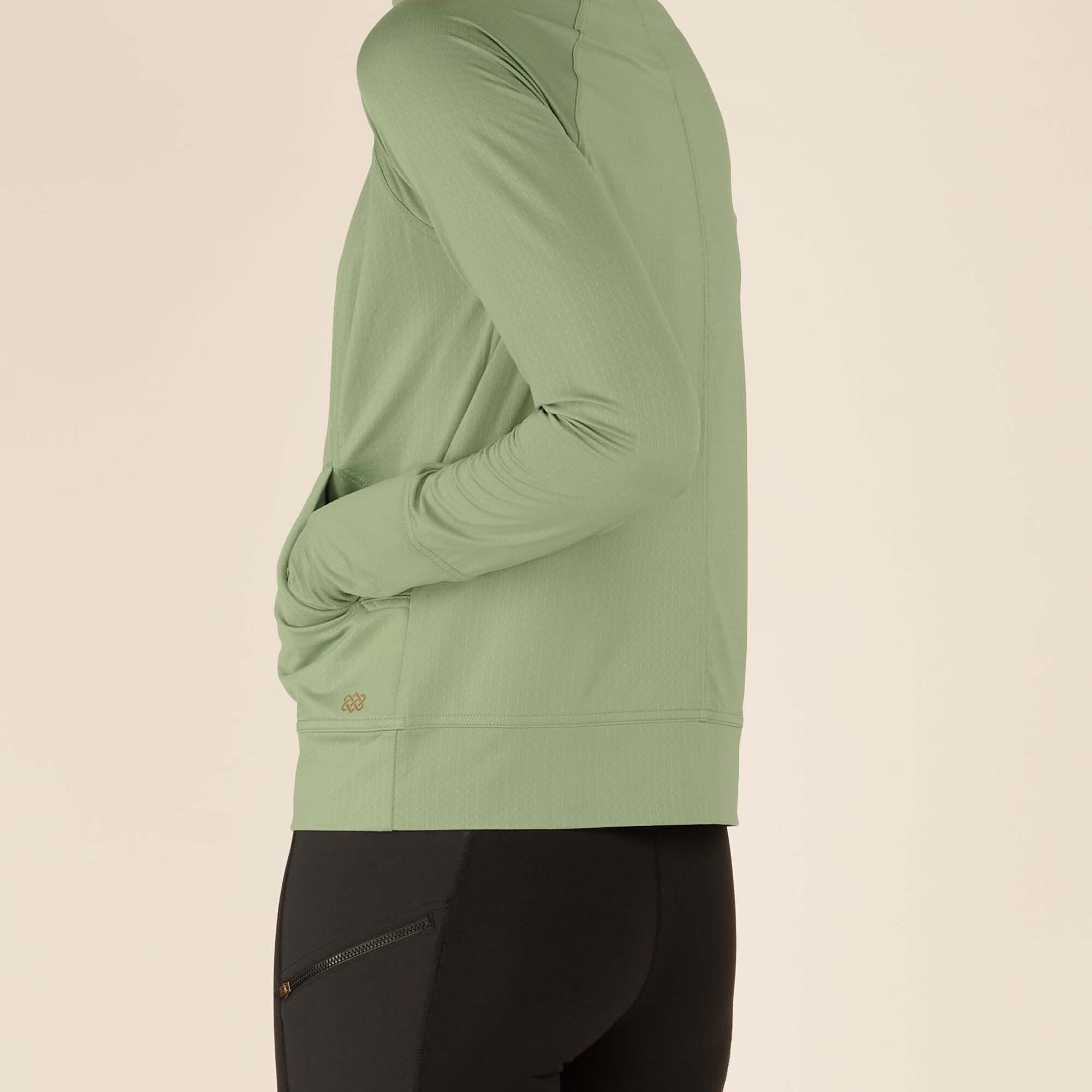 A close-up view of the lower portion of the Sherpa Adventure Gear Aarti Full Zip Jacket in Green, highlighting the side pocket detail with the Sherpa Adventure Gear logo subtly printed in gold near the hem. The fabric has a subtle texture, and the fit contours closely to the model’s body. This close-up also shows the thumbhole cuff design.