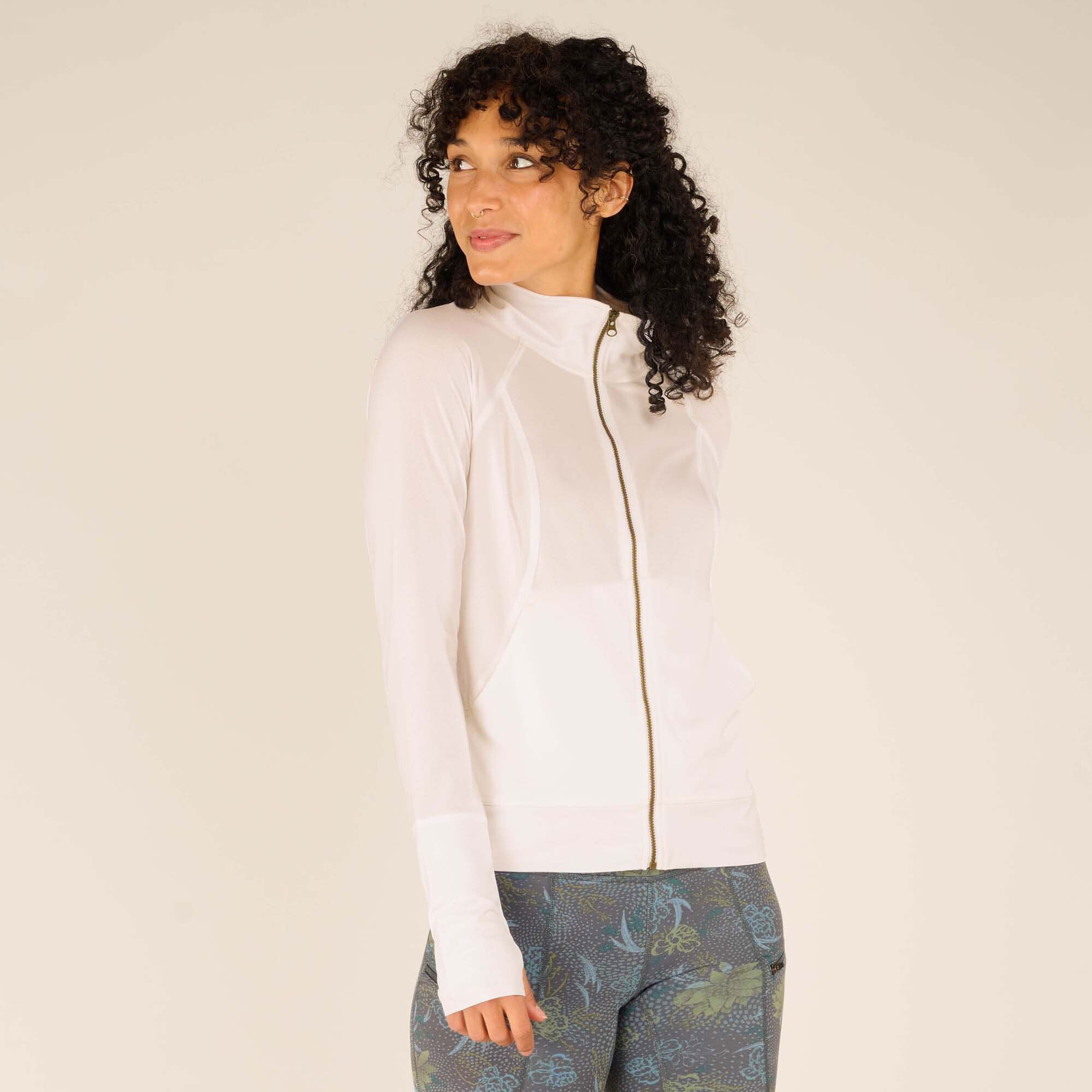 A woman with curly black hair and a medium skin tone is modeling a Sherpa Adventure Gear Aarti Full Zip Jacket in White. She is turned slightly to her left, looking off to the side with a soft smile. The jacket features a high collar, thumbholes on the sleeves, and a front zipper. She pairs it with blue-green floral-patterned biker shorts. The background is a neutral beige.