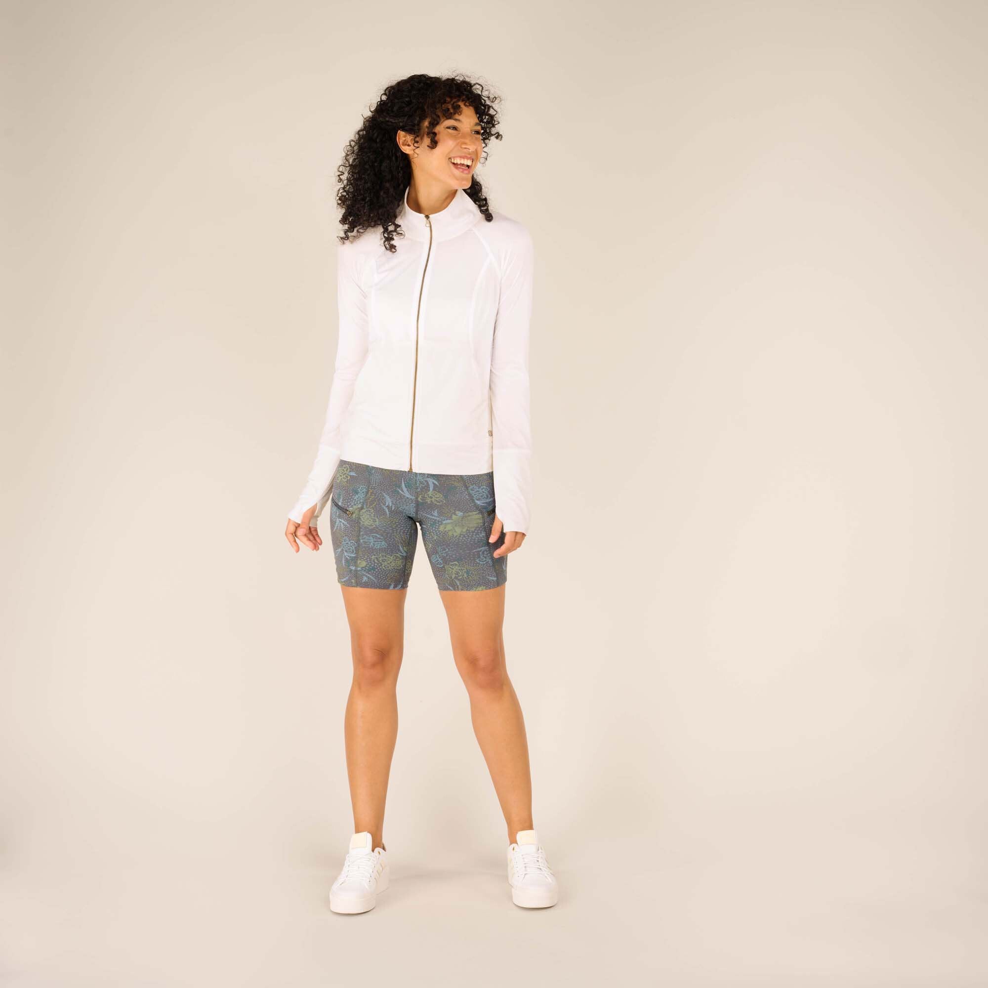 A full-body shot of the same woman standing confidently with her feet slightly apart, wearing the Sherpa Adventure Gear Aarti Full Zip Jacket in White and floral-patterned biker shorts. She is smiling broadly, with her hands at her sides. She pairs the outfit with white sneakers.