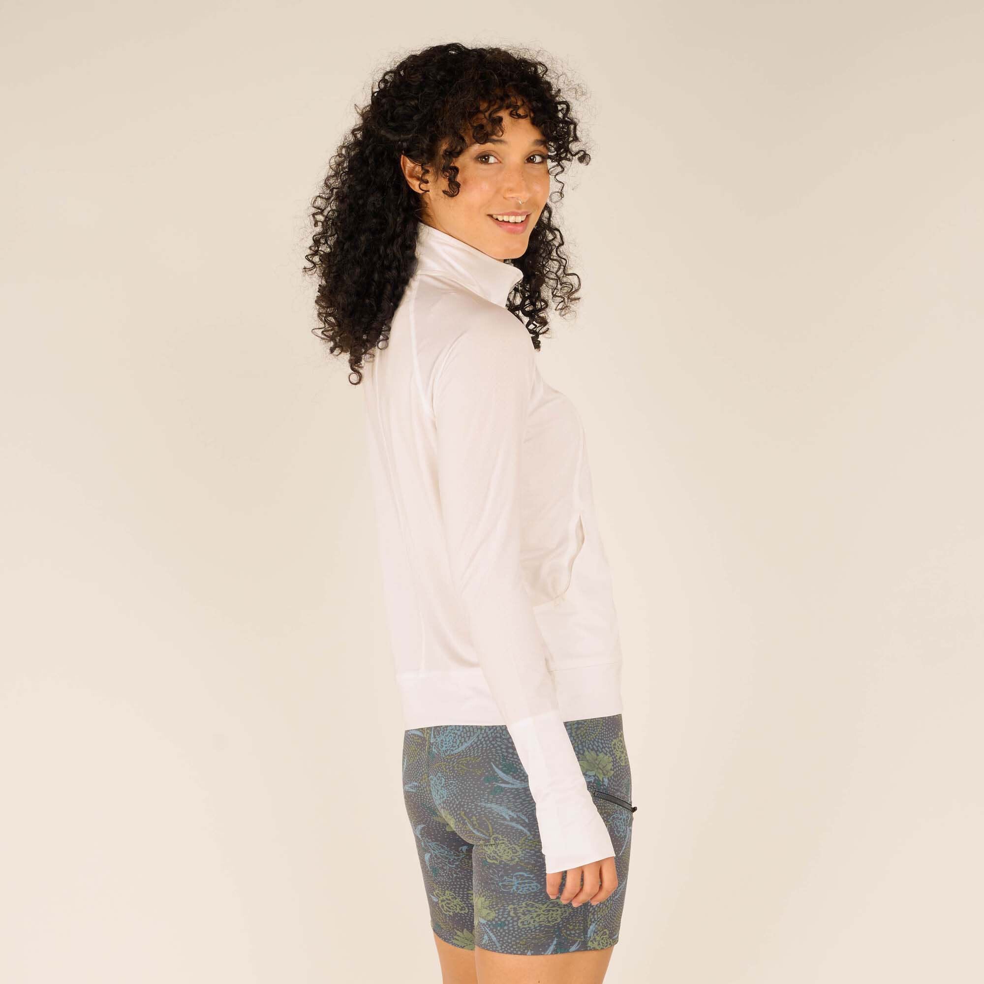 A side profile of the woman modeling the Sherpa Adventure Gear Aarti Full Zip Jacket in White, turned slightly away from the camera. She has a relaxed expression, with her curly hair framing her face. The back and side seams of the jacket are visible, along with the high collar and fitted silhouette.