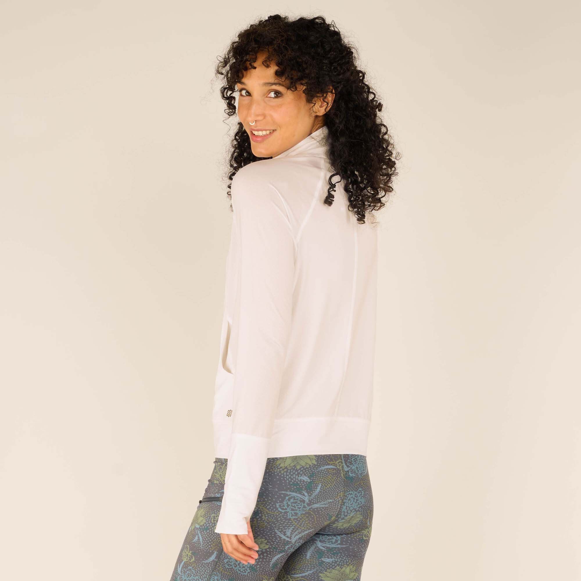 A three-quarter rear view of the woman, looking over her shoulder and smiling. The back details of the Sherpa Adventure Gear Aarti Full Zip Jacket in White, including the seams and fit, are visible. The shorts are also in view, showing their floral pattern and snug fit.