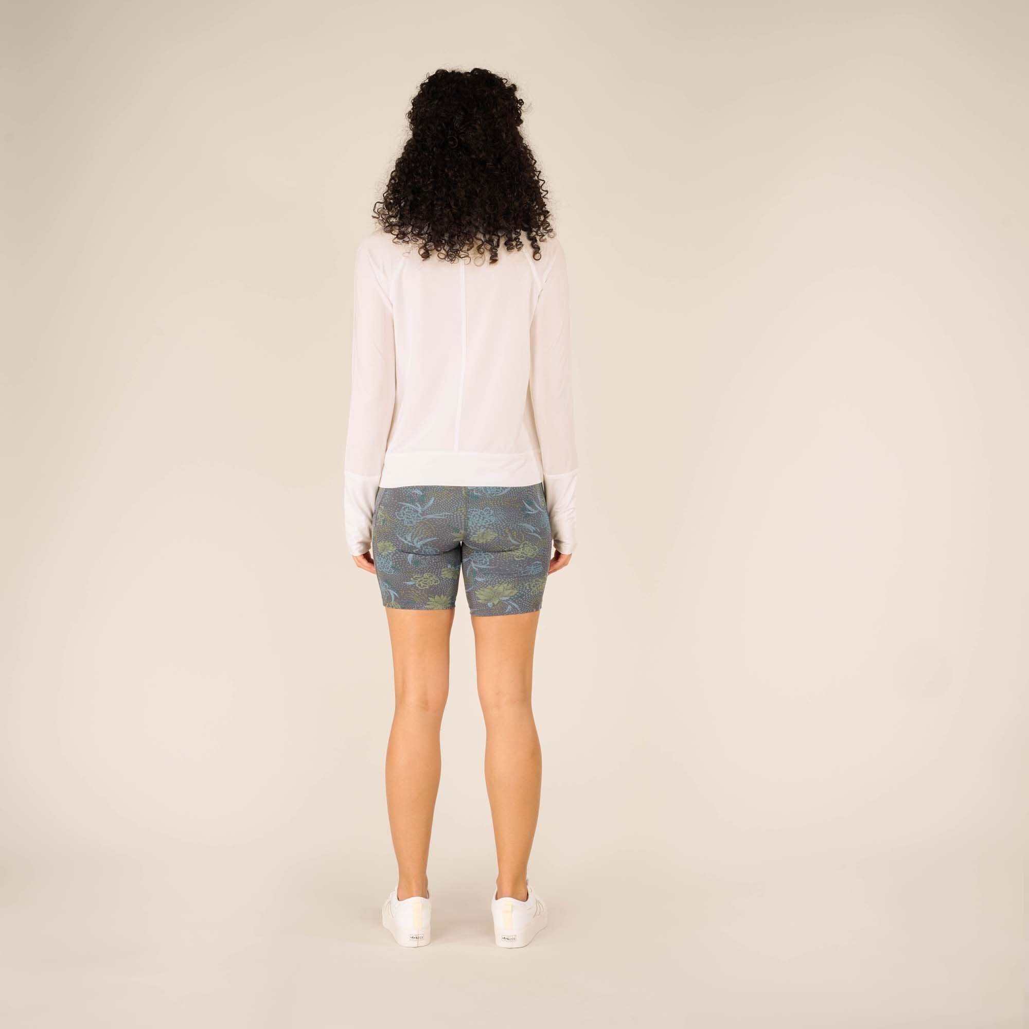 A rear full-body shot of the model with her back facing the camera, displaying the entire back of the Sherpa Adventure Gear Aarti Full Zip Jacket in White and shorts. She stands straight with her hands at her sides, and her curly hair cascades down her back.