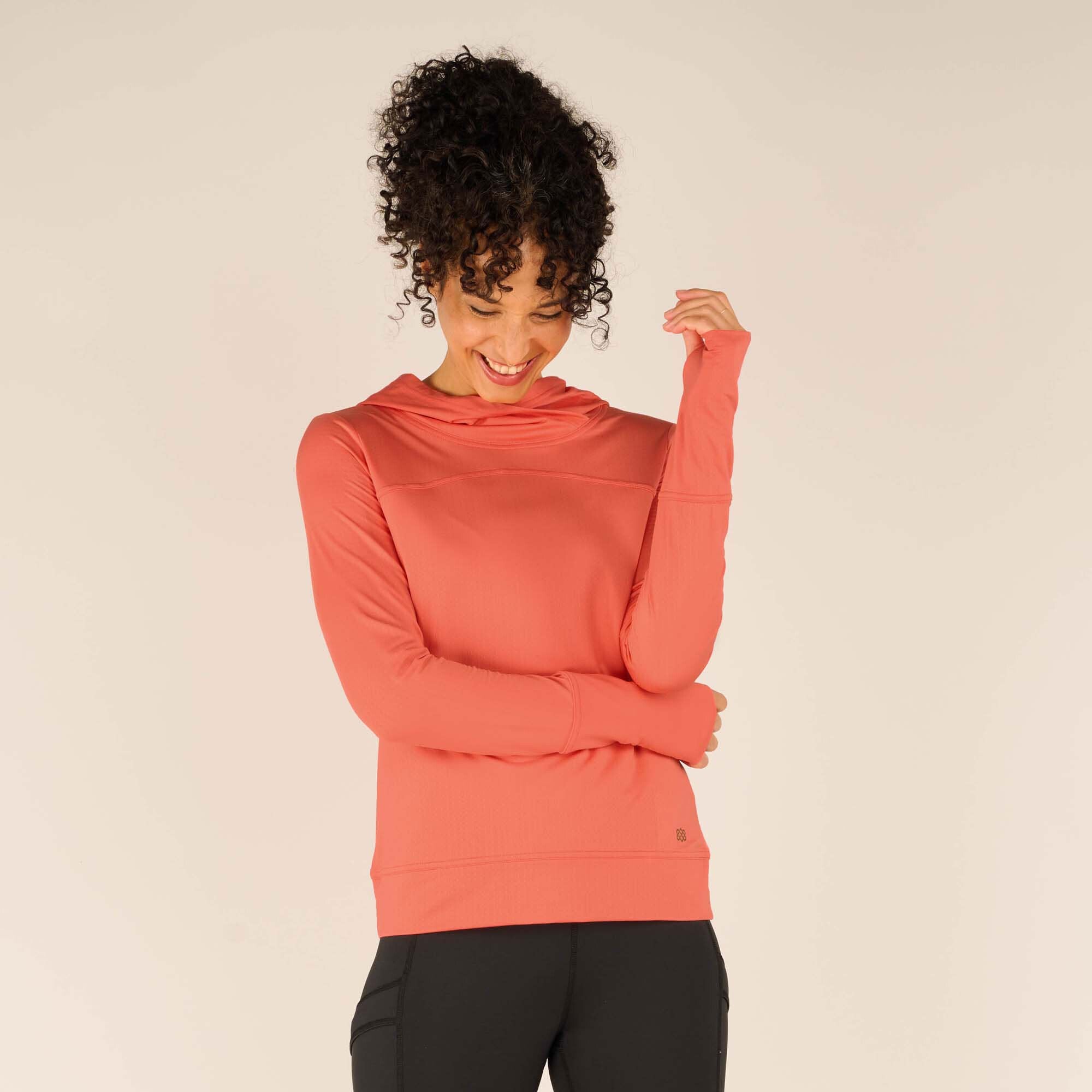 A woman with curly hair wears a Sherpa Adventure Gear Aarti Hoodie in Pink with a high neckline and a relaxed fit. She stands against a plain beige background, looking down and smiling, with her right arm crossed over her body and her left hand playfully touching her hair. The fabric appears soft and lightweight.