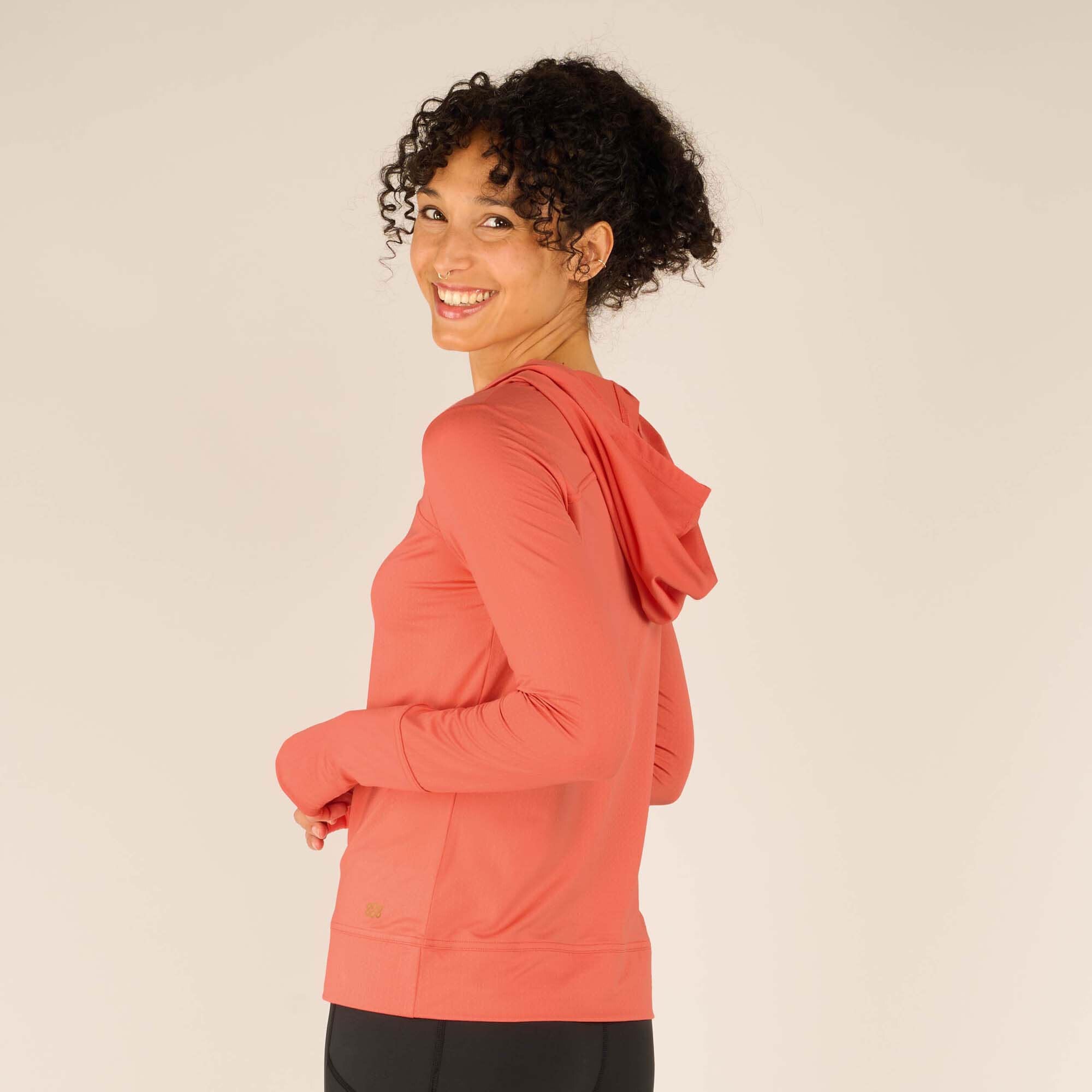 Three-quarter back view of the woman, turning her head to smile over her shoulder. The hood is visible, and subtle stitching detail runs horizontally across the upper back. The Sherpa Adventure Gear Aarti Hoodie in Pink maintains its relaxed fit.