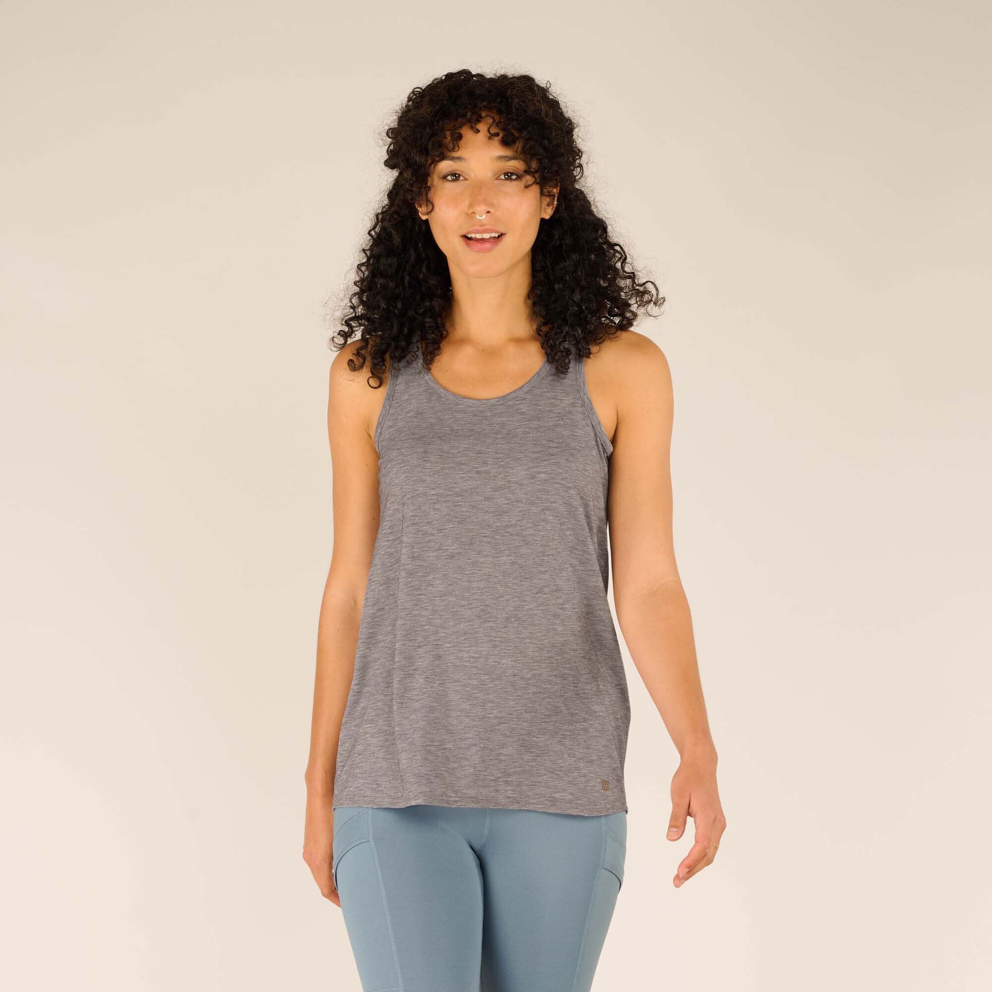 A woman is wearing a Sherpa Adventure Gear Asha Tank in Grey with a rounded neckline and relaxed fit. She is standing upright, facing forward, paired with light blue leggings and white trainers. The tank top has a subtle logo near the hemline.