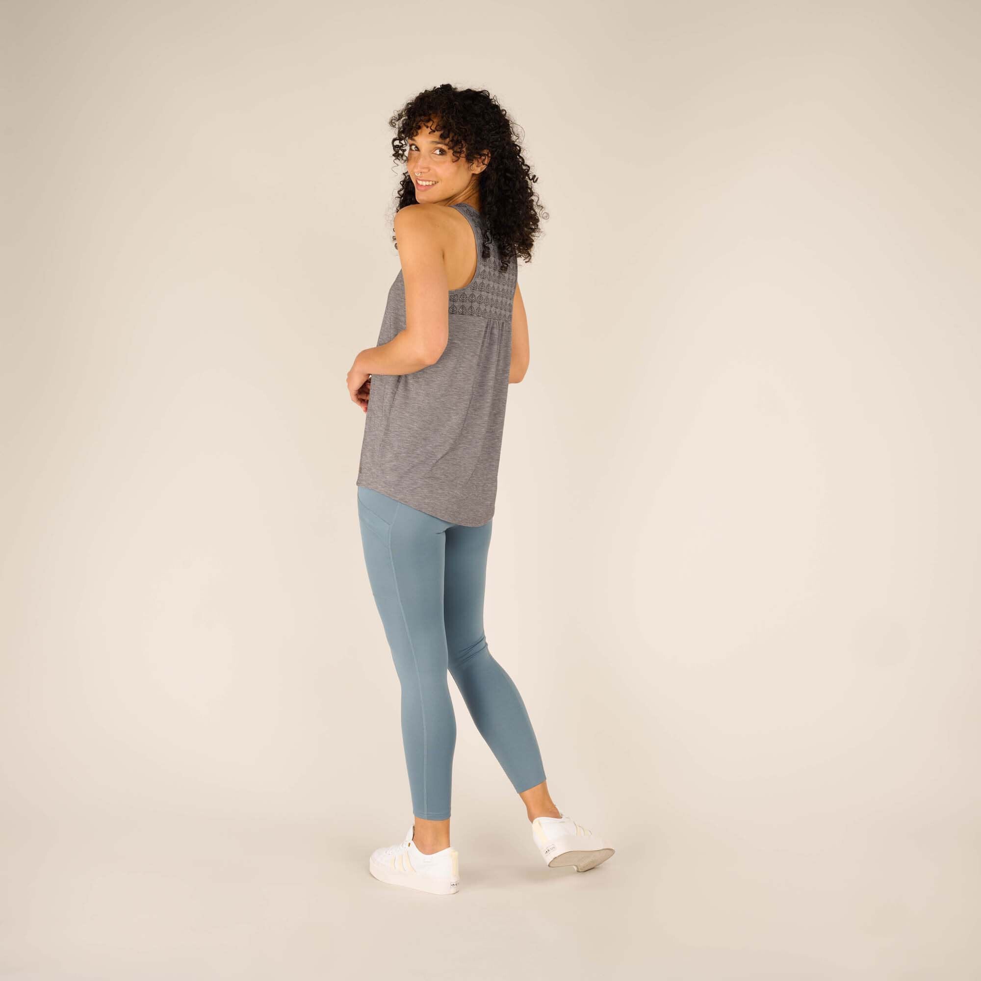 A side view of the same woman wearing the Sherpa Adventure Gear Asha Tank in Grey. The back features a patterned yoke design, showcasing small leaf motifs. The loose fit extends slightly below the hips, complementing her light blue leggings and white trainers.