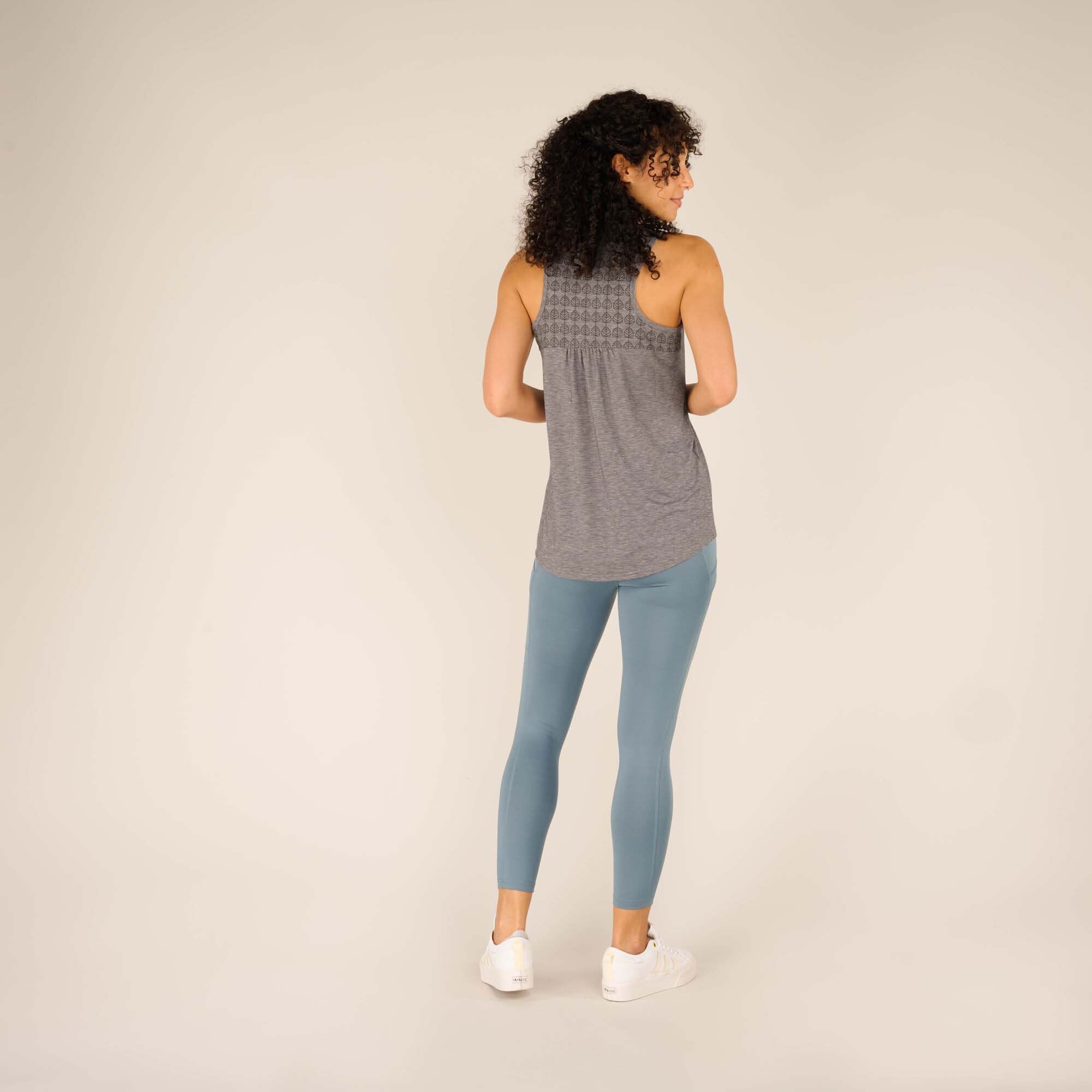 A full-back view of the woman wearing the Sherpa Adventure Gear Asha Tank in Grey. The patterned yoke design is prominently displayed at the upper back, and the rest of the fabric flows freely. She pairs it with light blue leggings, standing in a neutral pose.