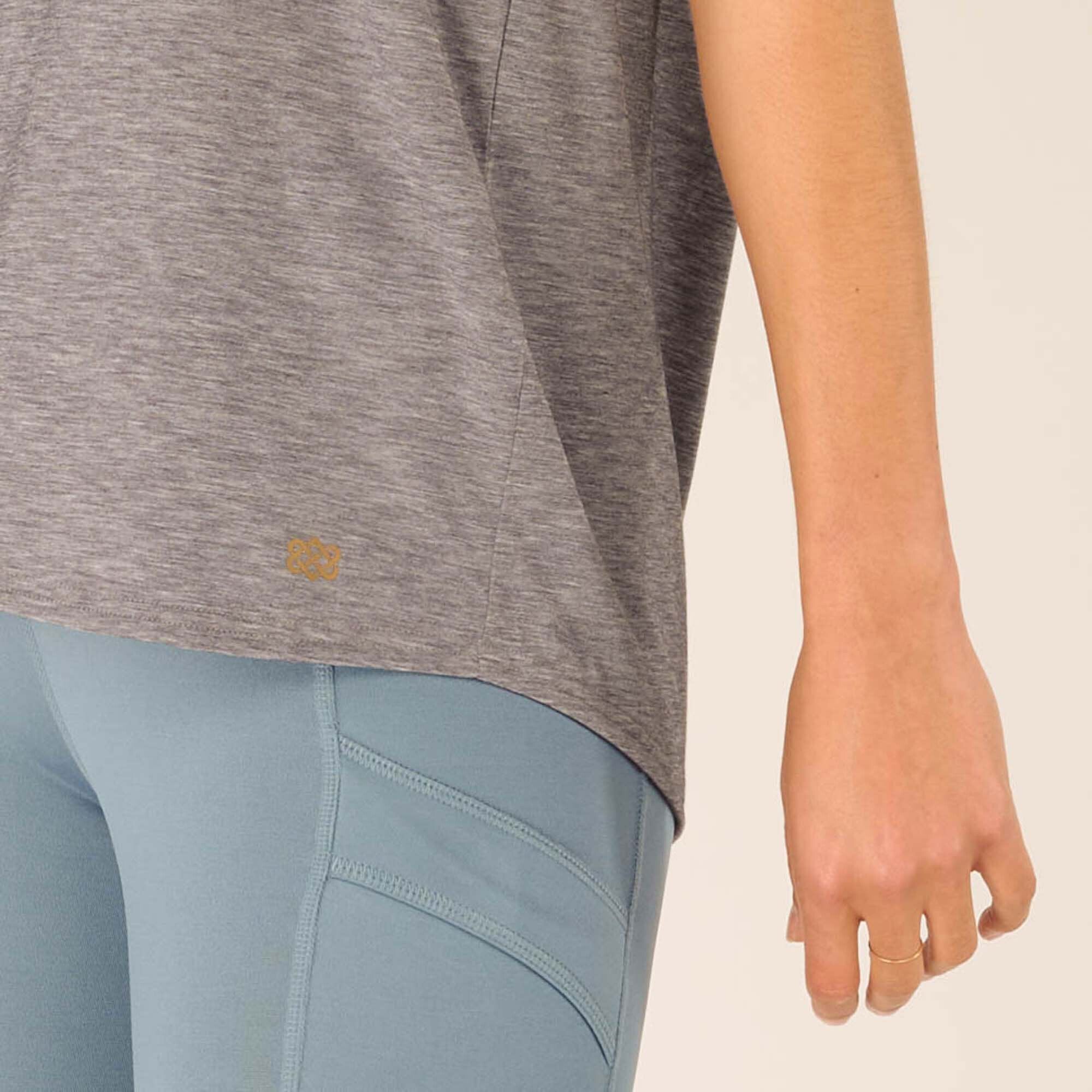 A close-up of the lower hem of the Sherpa Adventure Gear Asha Tank in Grey, highlighting the small golden Sherpa logo. The hemline is slightly curved and paired with blue leggings that feature visible seam details.