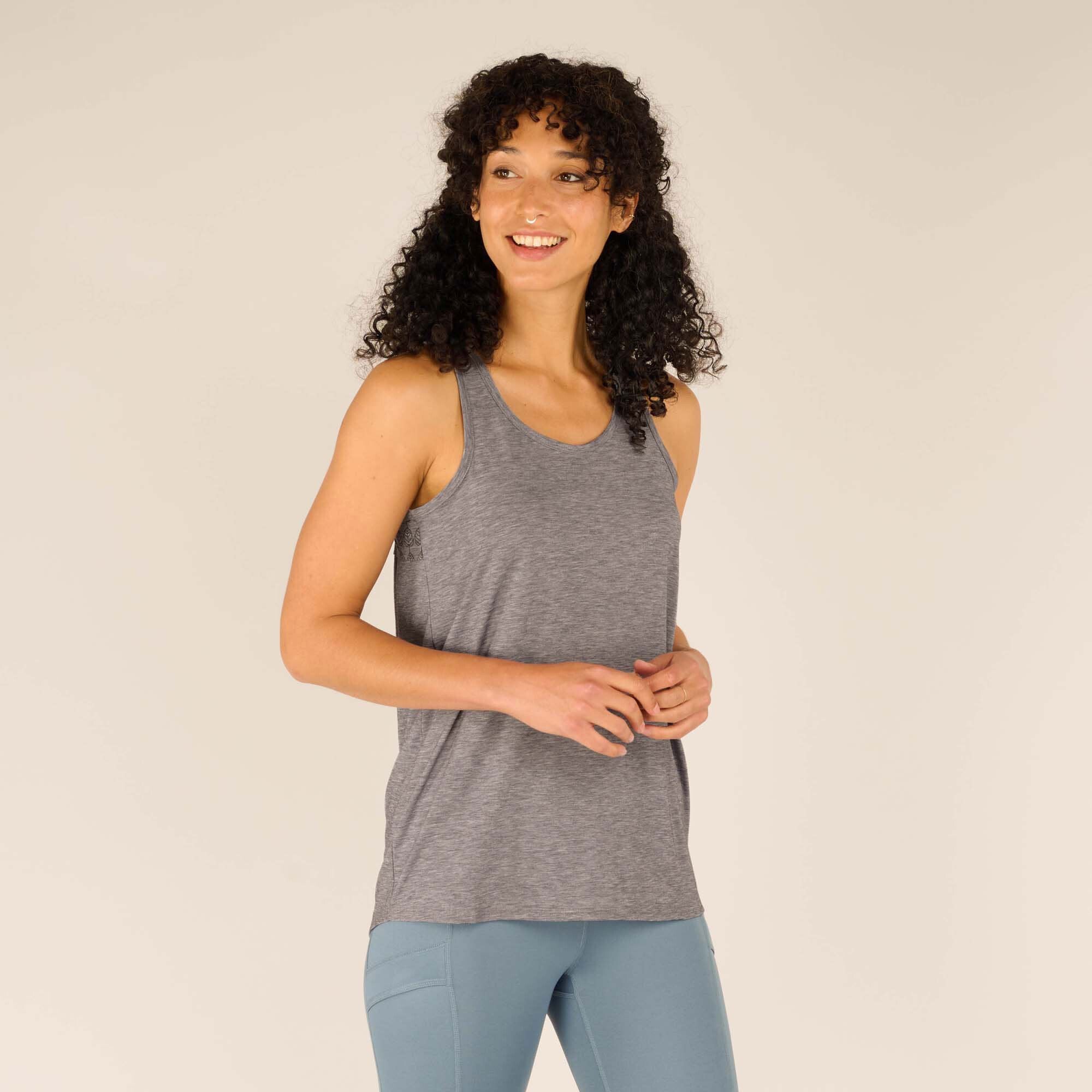 A forward-facing view of the woman wearing the Sherpa Adventure Gear Asha Tank in Grey. She is smiling, showcasing the relaxed fit and sleeveless design, complemented by light blue leggings. The minimalist design emphasises versatility and comfort.