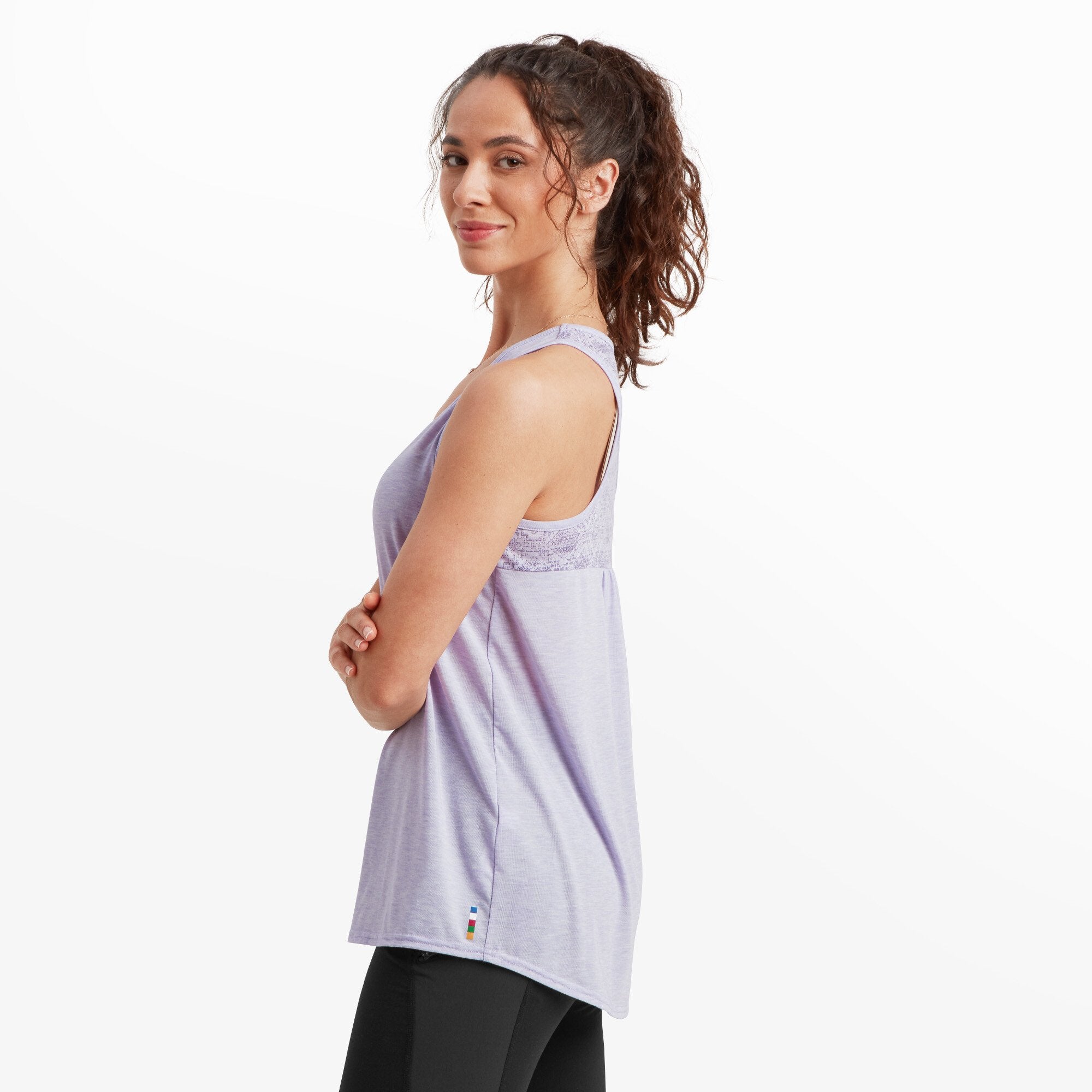 A side profile view of the woman wearing the Sherpa Adventure Gear Asha Tank in Purple. The back of the top showcases a patterned yoke in a light purple geometric design. The tank flows loosely over black leggings, and the woman stands with her arms crossed, looking towards the camera.