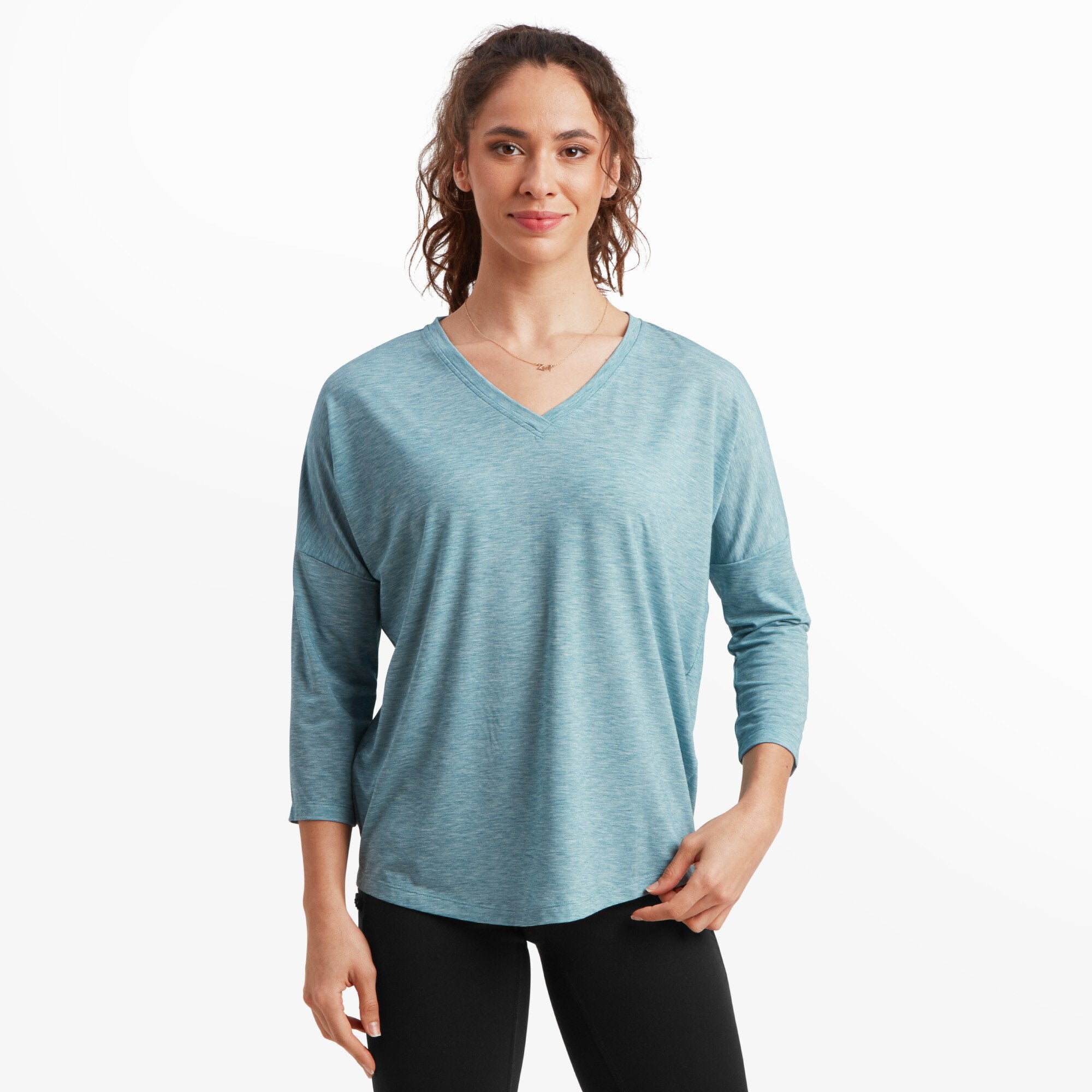 A woman with light skin and wavy brown hair tied in a loose ponytail is modeling a Sherpa Adventure Gear Asha V-Neck 3/4 Sleeve Top in Blue. She is standing with a neutral expression and soft smile against a white background. The fabric appears lightweight, and she is also wearing black leggings.