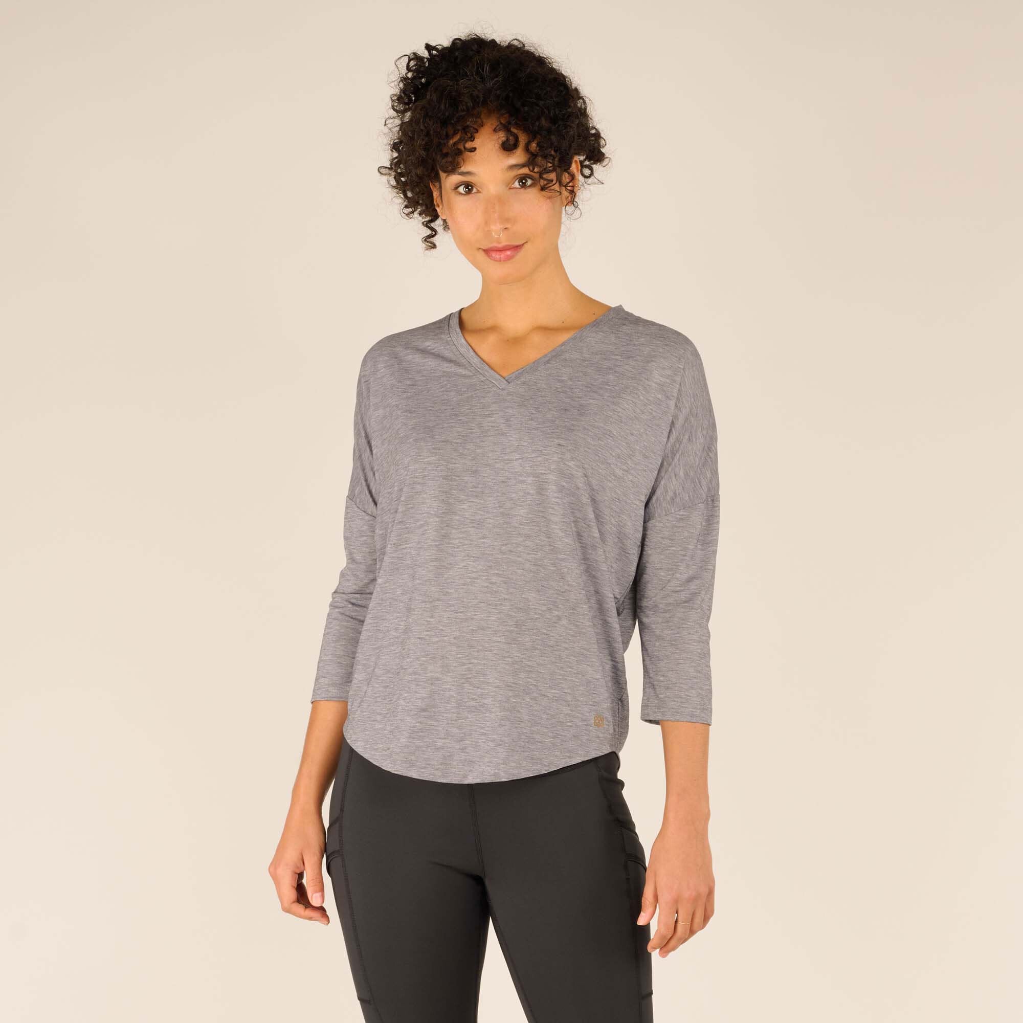 A woman is wearing a relaxed-fit, Sherpa Adventure Gear Asha V-Neck 3/4 Sleeve Top in Grey with a V-neckline and 3/4 length sleeves. The fabric appears soft and breathable, and the top is paired with black leggings for a casual, comfortable look. The model is posing confidently against a light beige background.