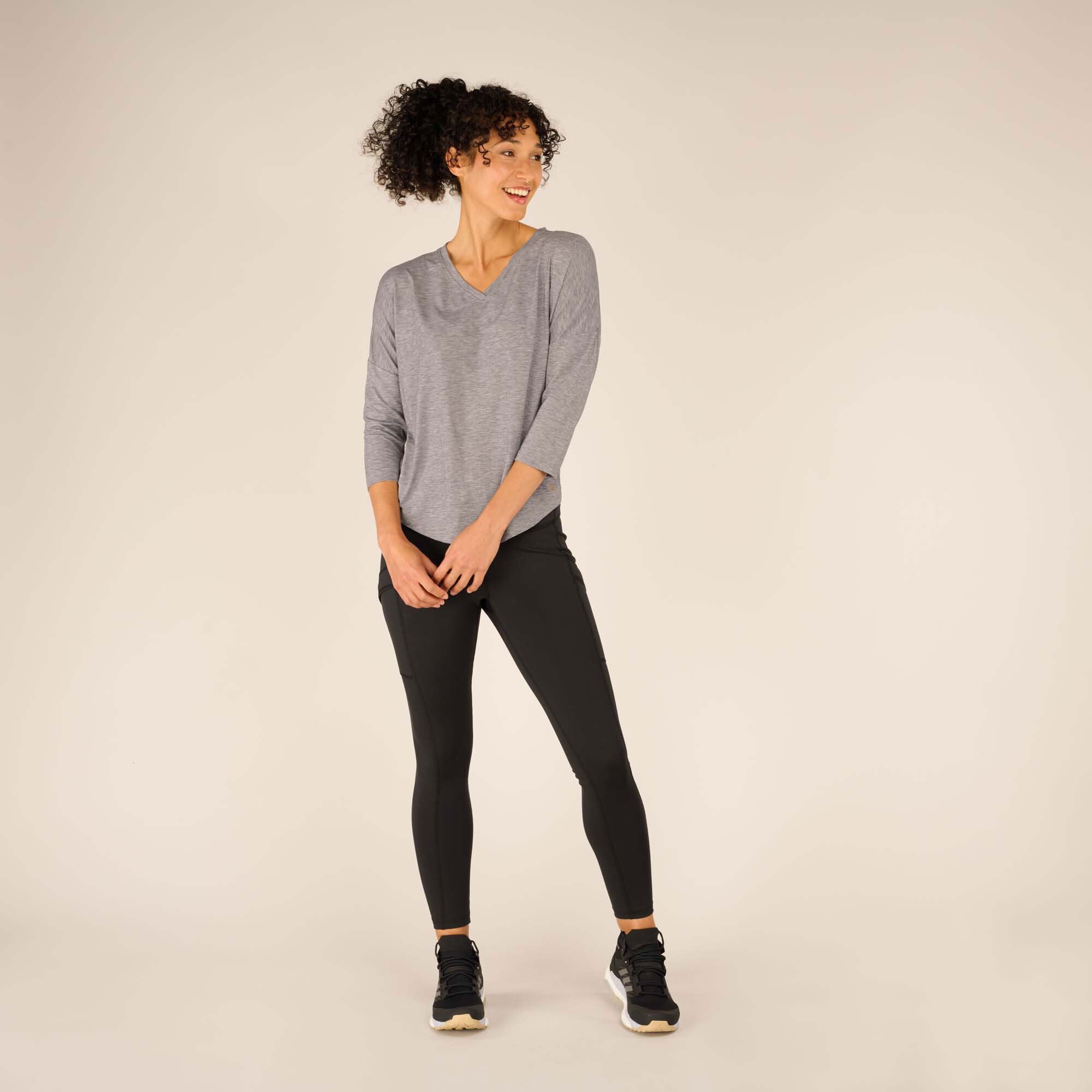 The same woman is shown standing with a playful, casual stance. The Sherpa Adventure Gear Asha V-Neck 3/4 Sleeve Top in Grey drapes naturally on her frame, while the black leggings and black trainers complete her sporty and relaxed outfit. She is smiling, exuding an approachable and stylish vibe.