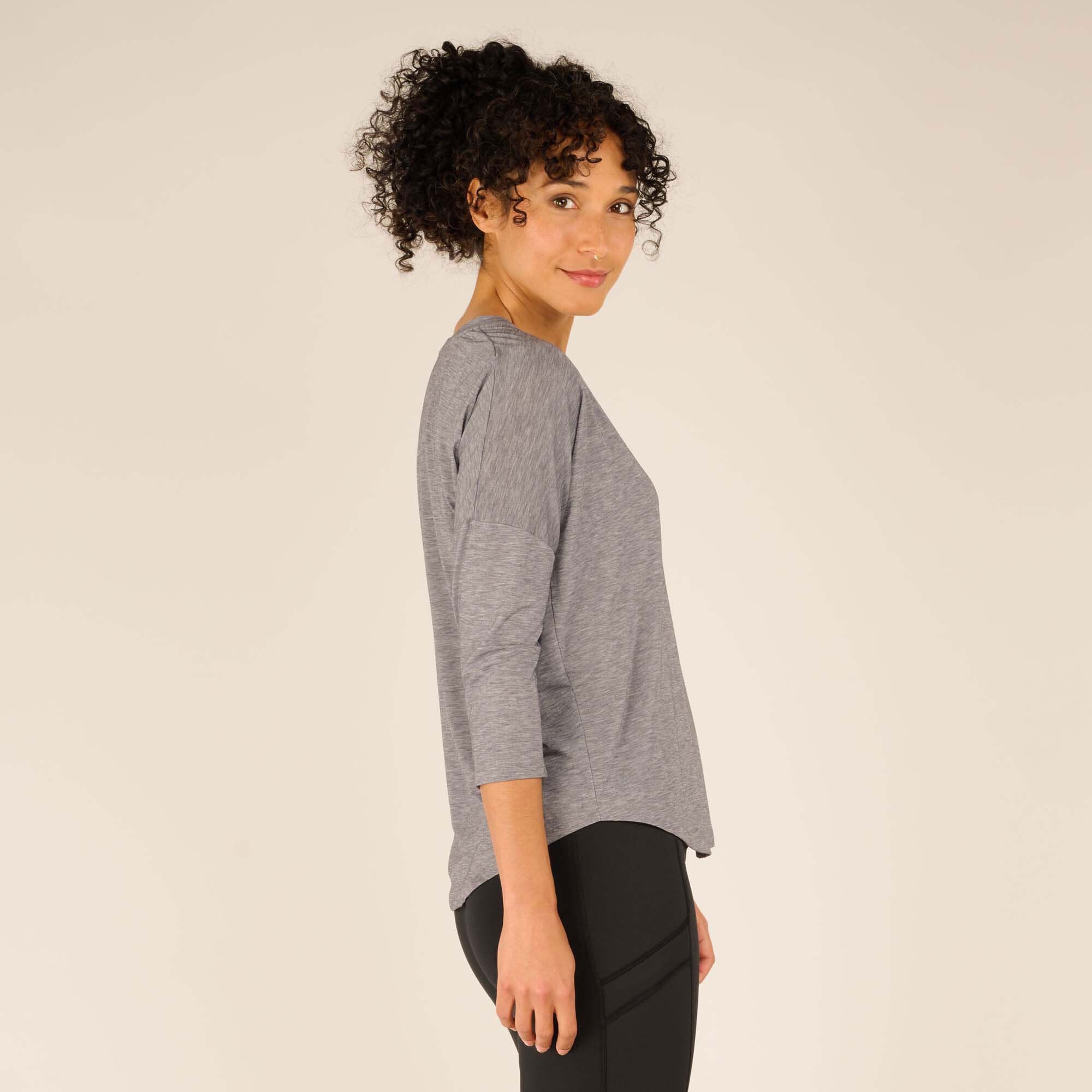 A side profile view of the woman highlights the loose fit of the Sherpa Adventure Gear Asha V-Neck 3/4 Sleeve Top in Grey. The top has dropped shoulders and gently flows past the hips, creating a flattering silhouette. Her curly hair is styled in a high ponytail, complementing the sporty aesthetic.