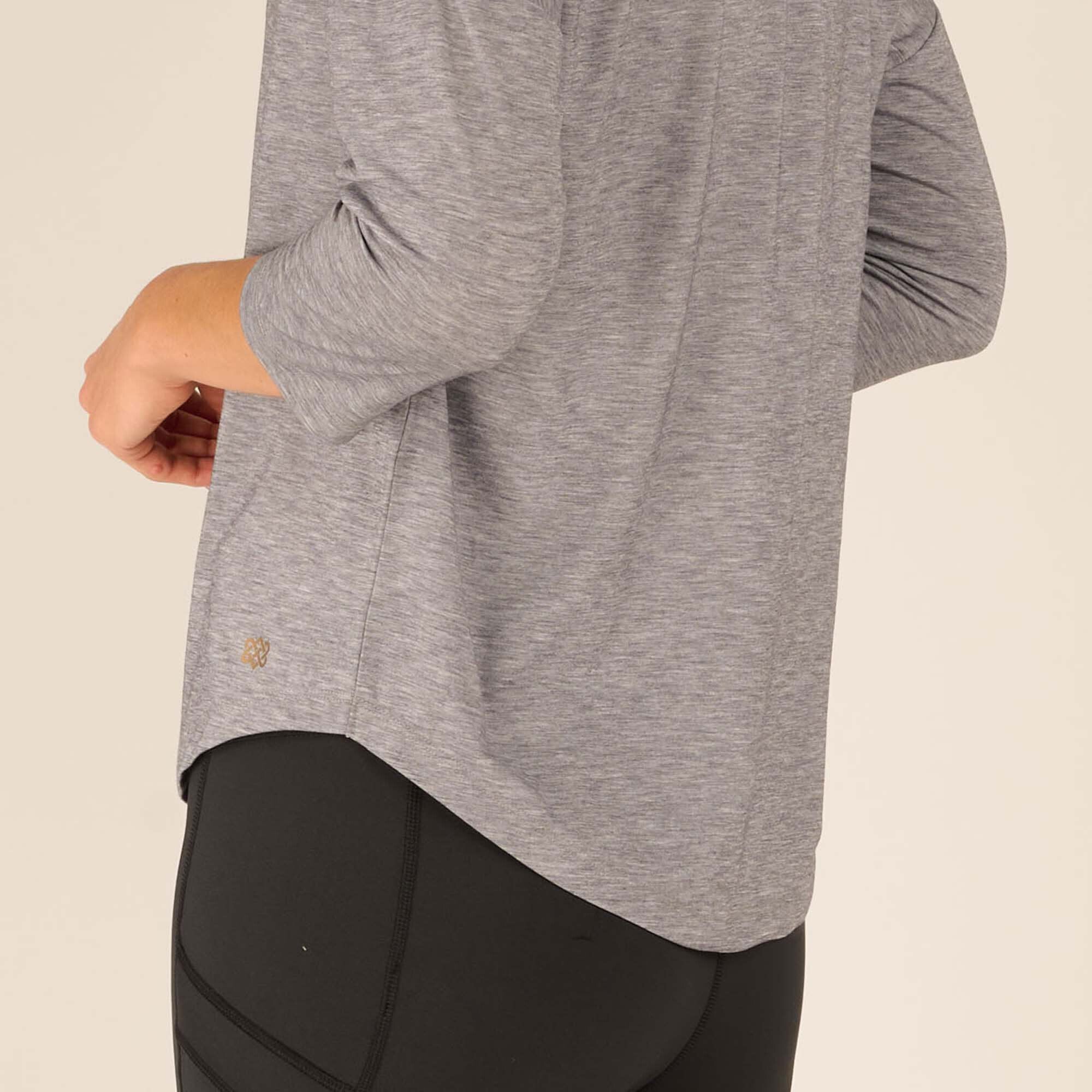 A close-up shot of the bottom hem of the Sherpa Adventure Gear Asha V-Neck 3/4 Sleeve Top in Grey reveals a small, subtle embroidered logo near the hemline. The fabric's soft texture and relaxed fit are evident, highlighting the garment's quality and design. The black leggings provide a sleek contrast to the grey top.