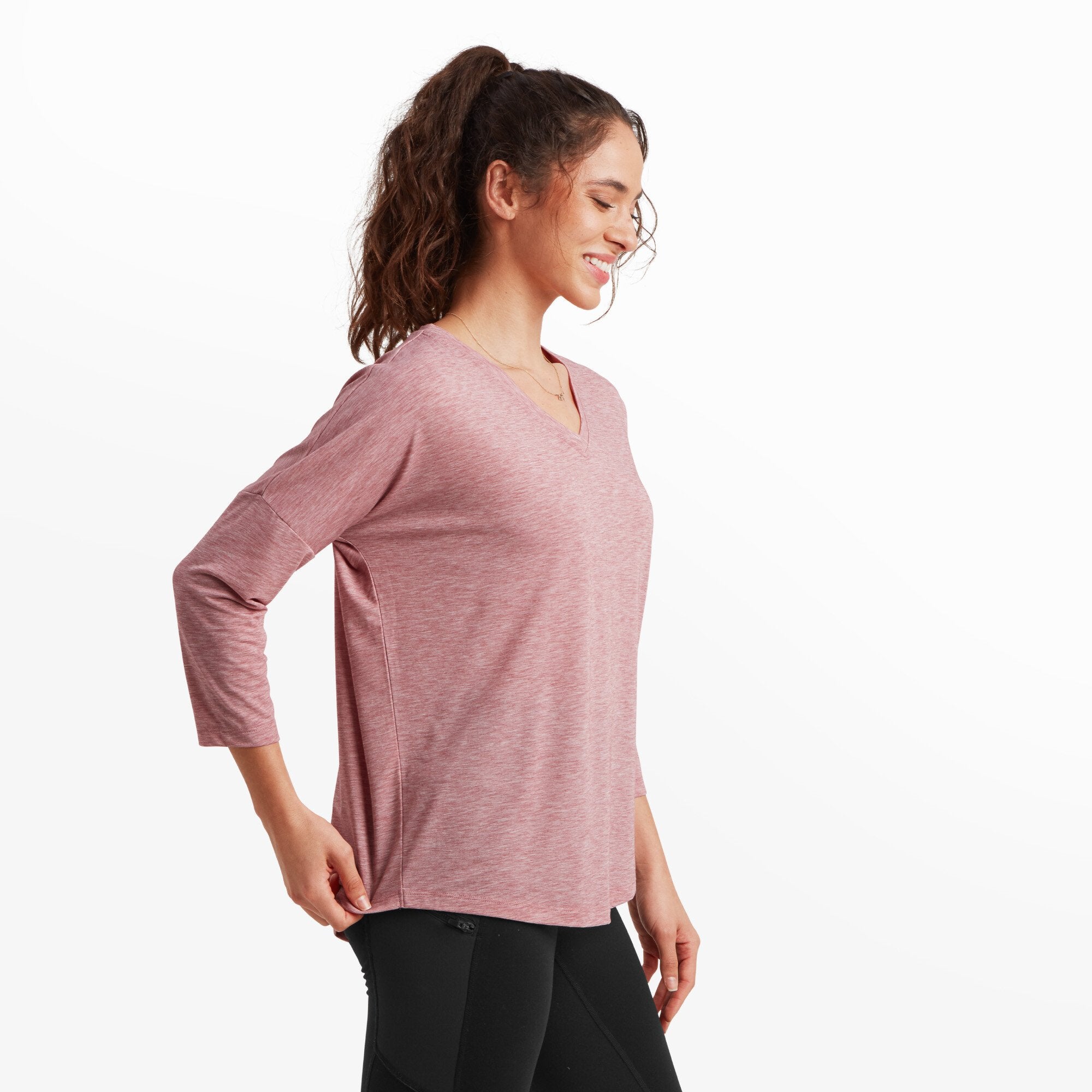 The same woman is shown from the side, turning slightly with her left arm bent to reveal the side seam of the Sherpa Adventure Gear Asha V-Neck 3/4 Sleeve Top in Red. The 3/4 sleeves fall just below her elbows, and the hem has a gentle curve, emphasising the flowy nature of the fabric.