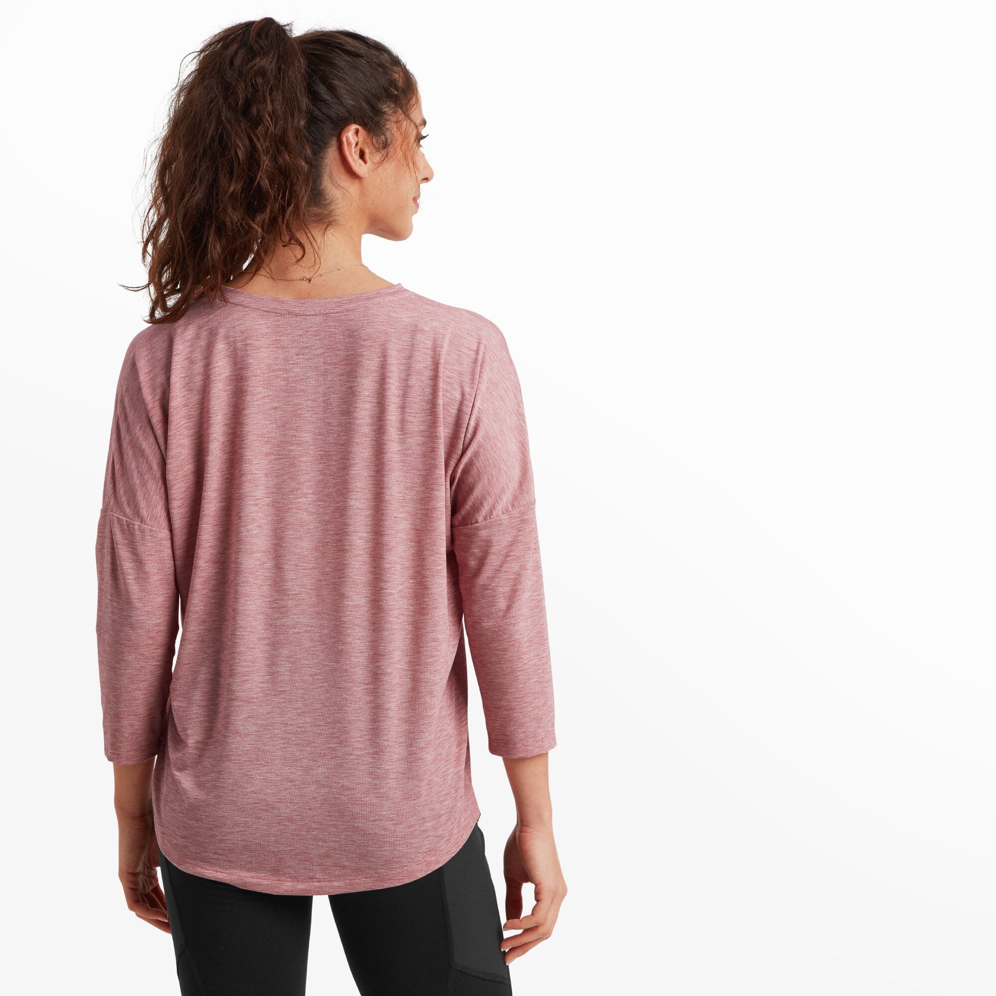 A back view of the Sherpa Adventure Gear Asha V-Neck 3/4 Sleeve Top in Red highlights the soft drape of the fabric on her shoulders and back. The top flows loosely, ending in a rounded hem that dips slightly longer in the back. Her ponytail and posture enhance the casual, everyday appeal of the shirt.