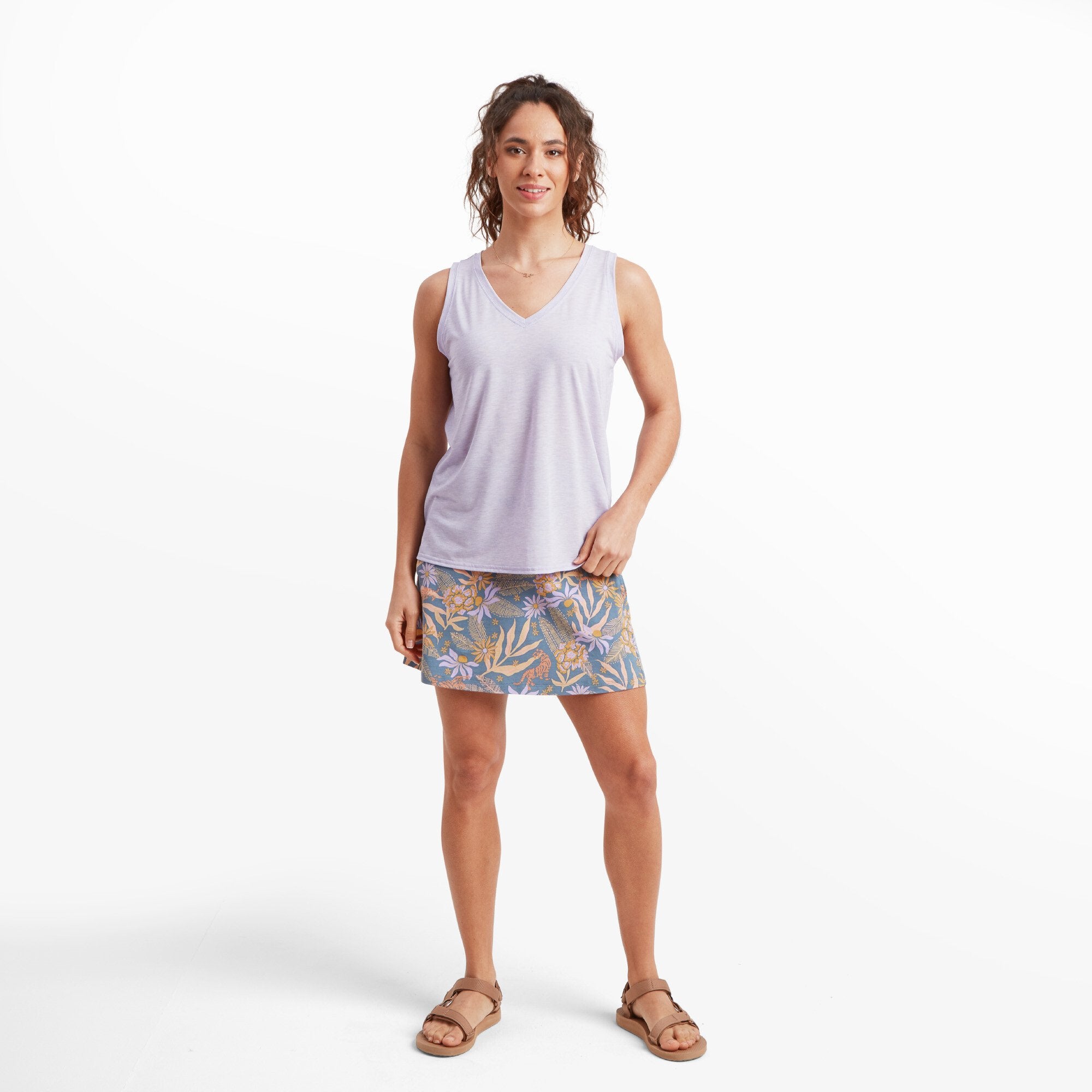 A full-body image of a woman wearing a Sherpa Adventure Gear Asha V-Neck Tank in Purple with a relaxed fit and a soft V-neckline. She pairs it with a patterned floral skirt and open-toed brown sandals, standing confidently with one hand resting on the skirt.