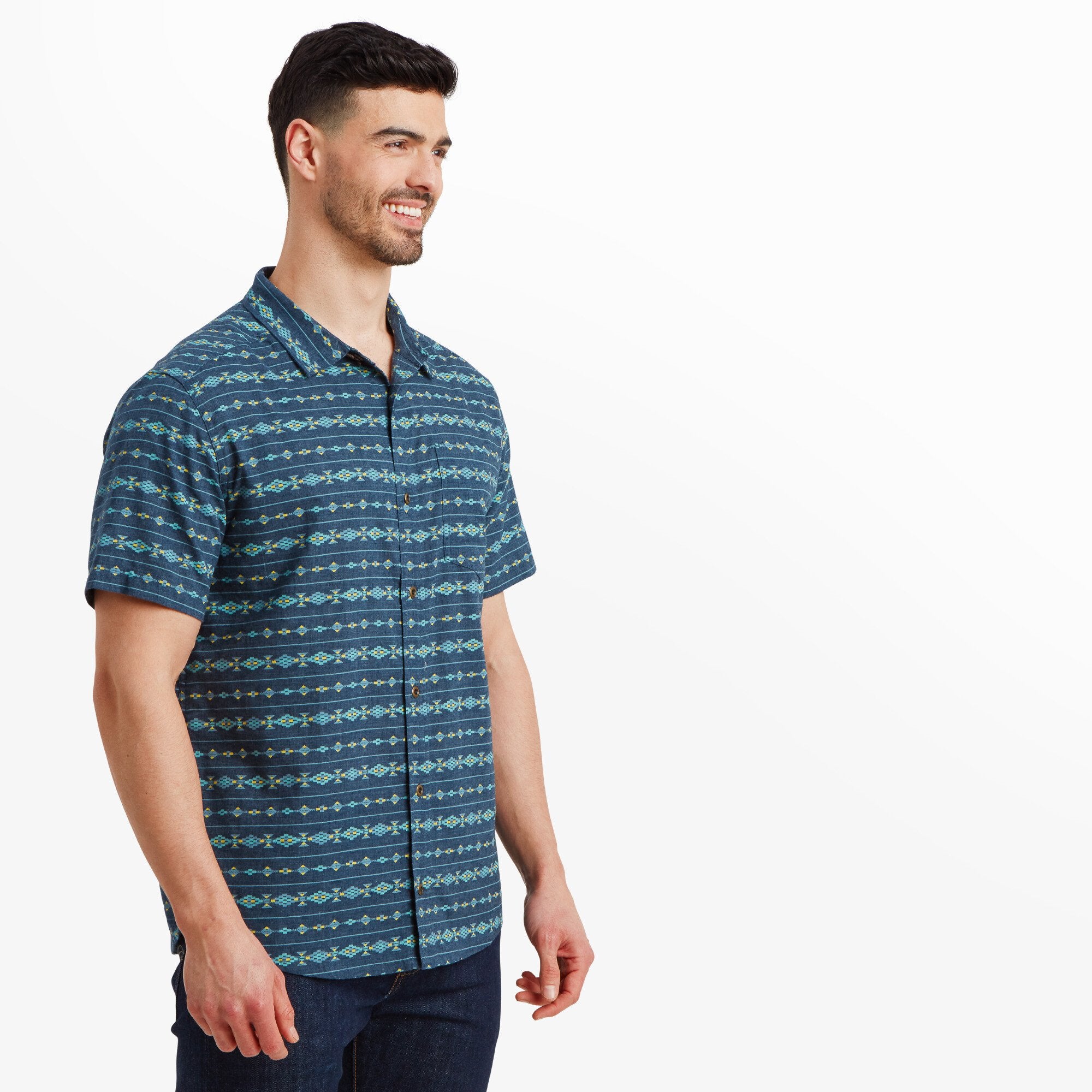 The model is standing with a slight smile, showcasing a Sherpa Adventure Gear Bagmati Stripe Short Sleeve Shirt in Blue featuring intricate horizontal geometric patterns in turquoise, yellow, and white. The shirt is paired with dark denim jeans, and the buttons are natural-toned wood.