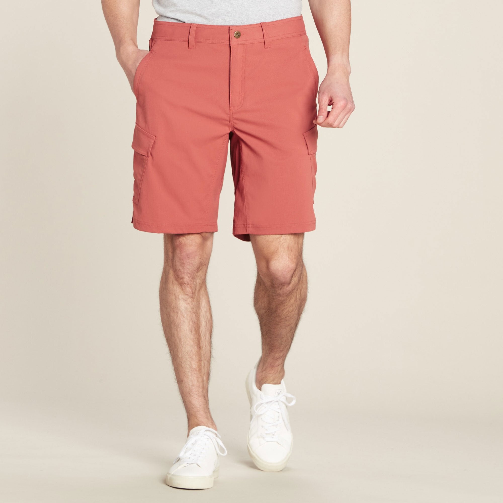 A male model wearing Sherpa Adventure Gear Bara Cargo Shorts in Beige, standing with his legs slightly apart. The shorts feature a straight fit above the knee with a button and zip closure. Two large cargo pockets are visible on the sides, and the model's hands are casually resting in the side pockets. The outfit is paired with white trainers, and the background is plain beige.