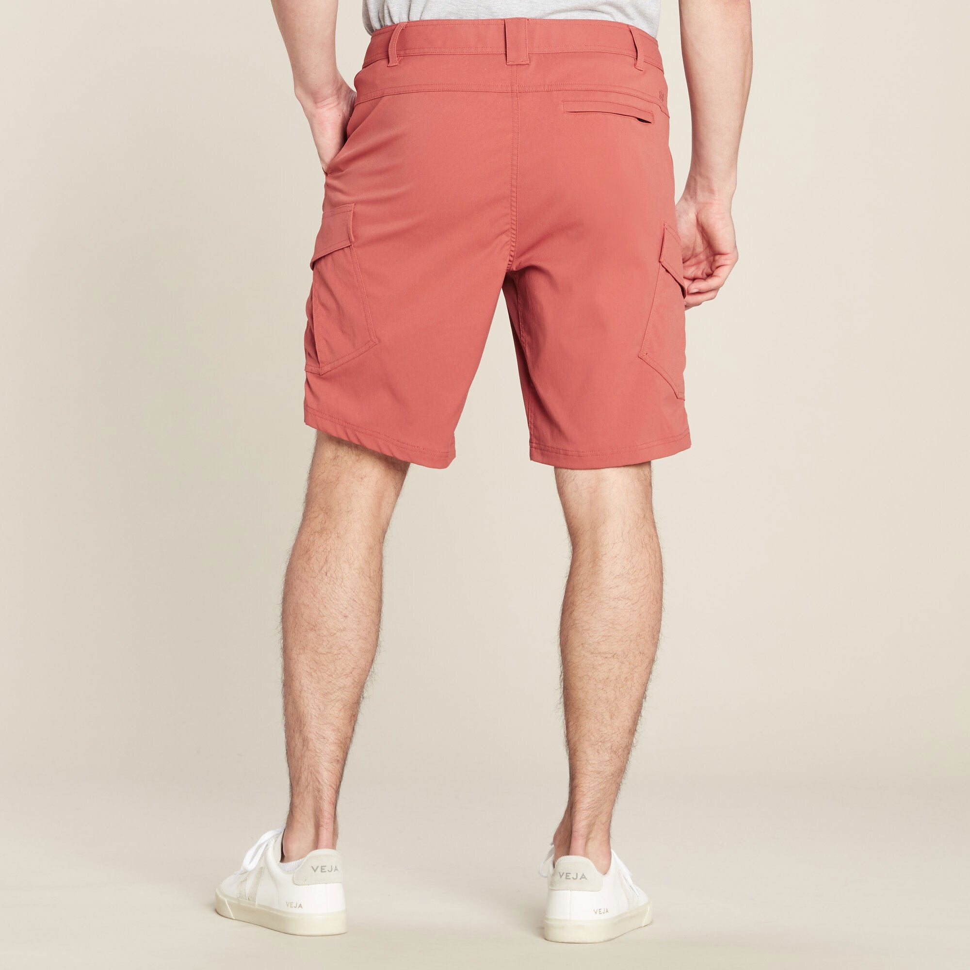 The back view of the Sherpa Adventure Gear Bara Cargo Shorts in Beige on the male model. The shorts have two back pockets—one with a flap and one with a clean finish. The model stands with a neutral pose, showing the mid-thigh length and the tailored fit of the shorts. The outfit is complemented by white trainers, and the background remains plain beige.