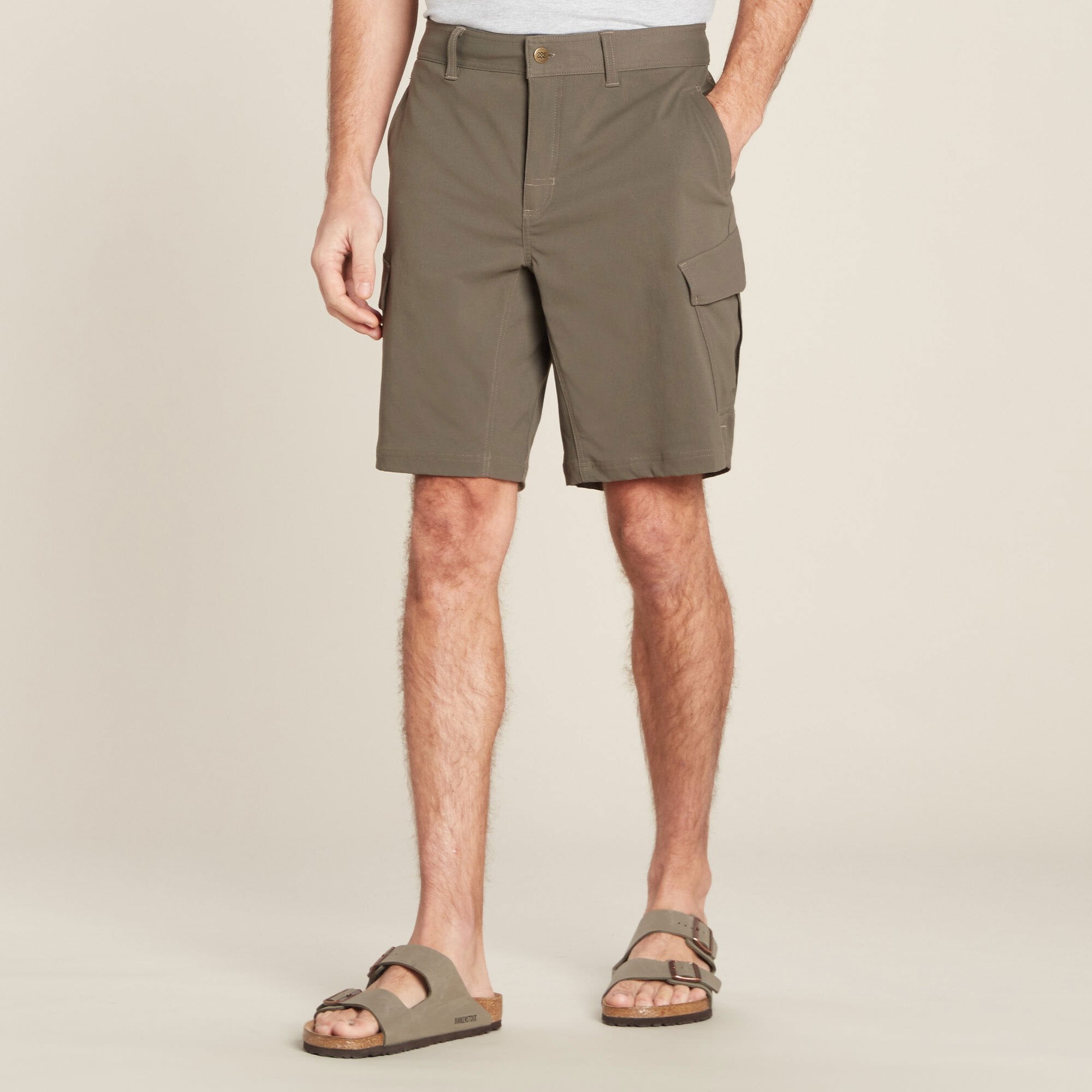 A front view of a man wearing Sherpa Adventure Gear Bara Cargo Shorts in Green paired with a gray t-shirt and beige sandals. The shorts feature a flat front with a button and zip closure, and a cargo pocket on the side. The man is standing upright with his left hand in the pocket, showcasing the relaxed fit of the shorts.