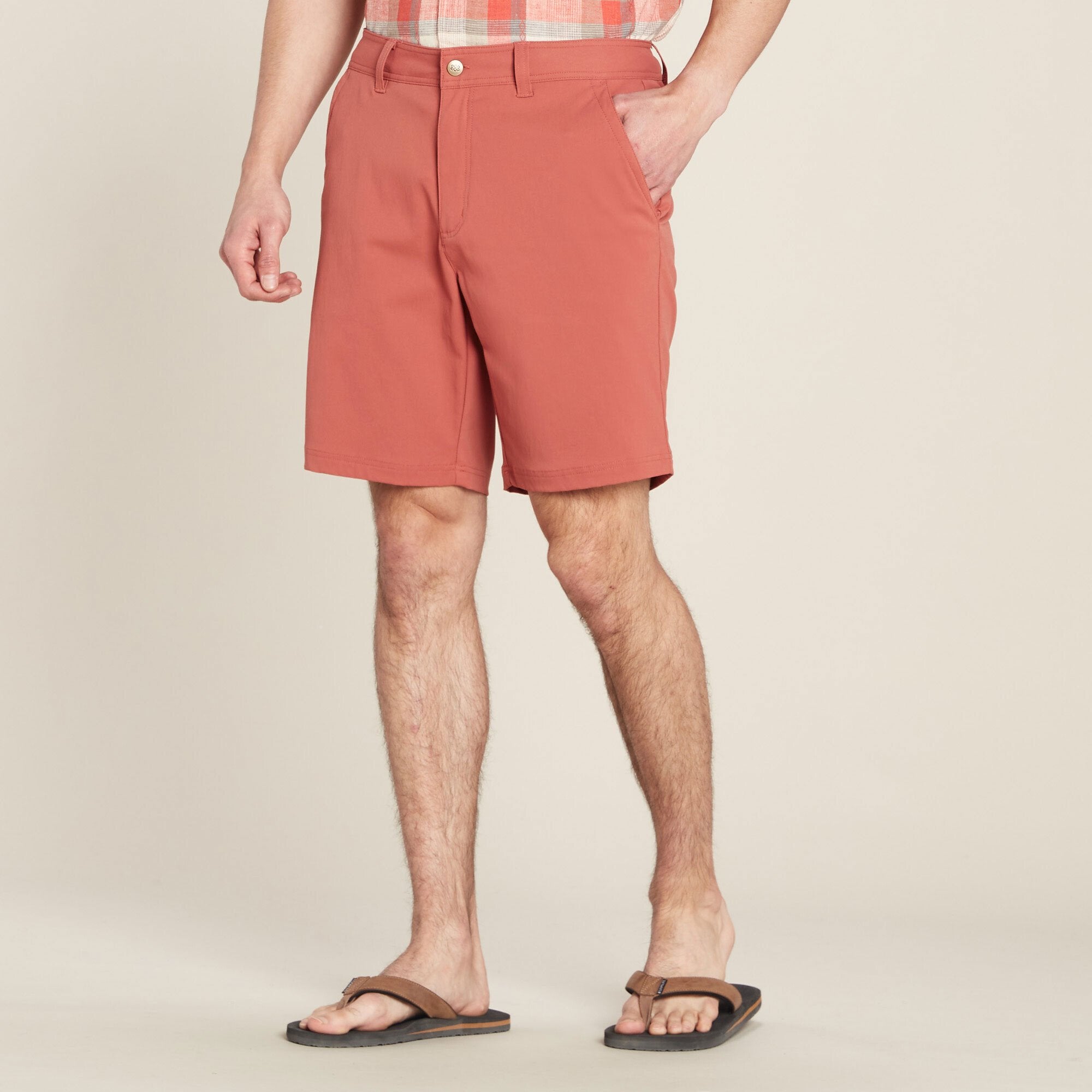 A close-up of a male model wearing Sherpa Adventure Gear Bara Shorts in Red paired with a plaid shirt, showcasing the front view of the garment. The shorts feature a clean, tailored design with side pockets, belt loops, and a button closure. The model is barefoot, standing on a neutral beige background.