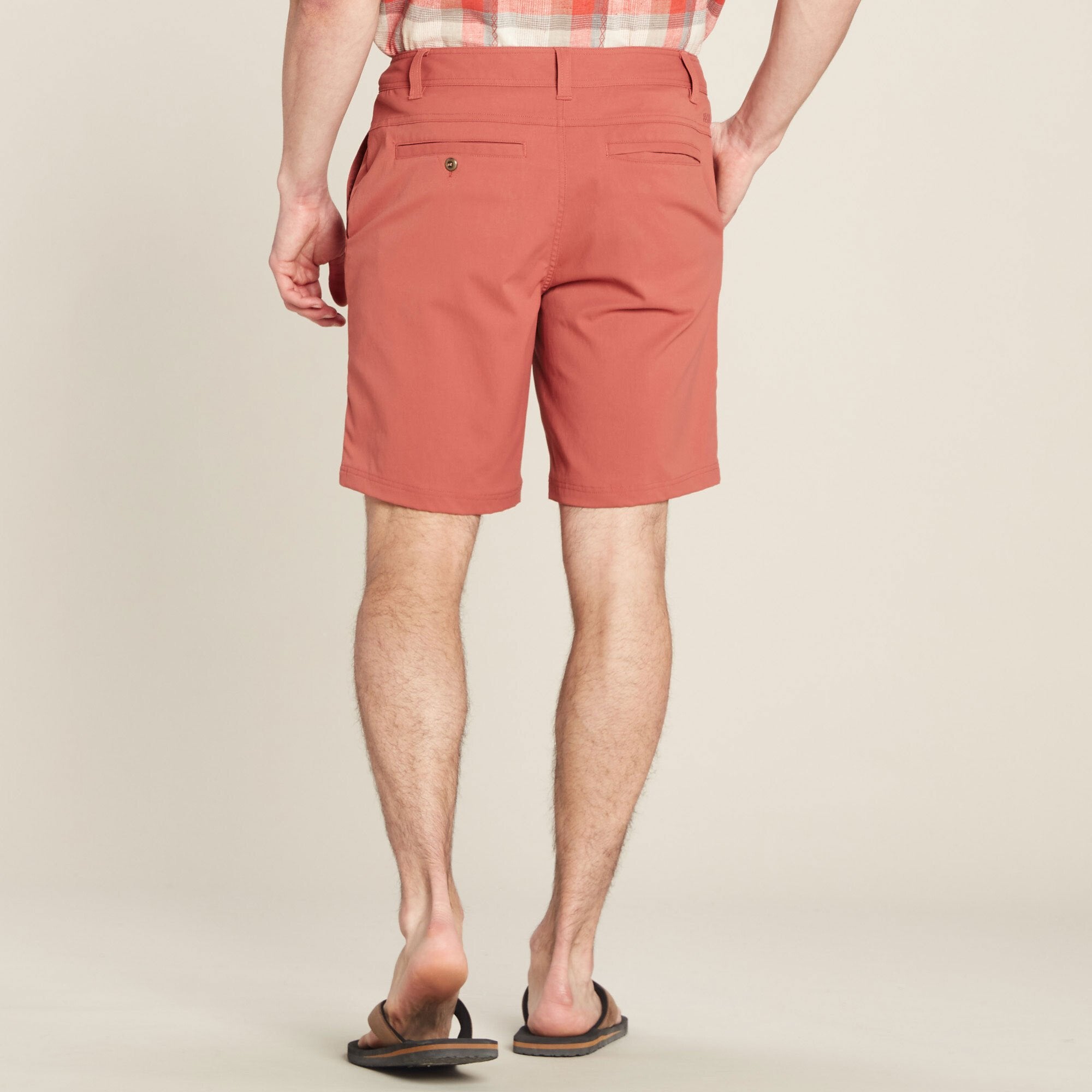 A rear view of a male model wearing Sherpa Adventure Gear Bara Shorts in Red. The shorts include back welt pockets, one with a button detail. The model's legs and sandals are visible, set against a simple beige background.