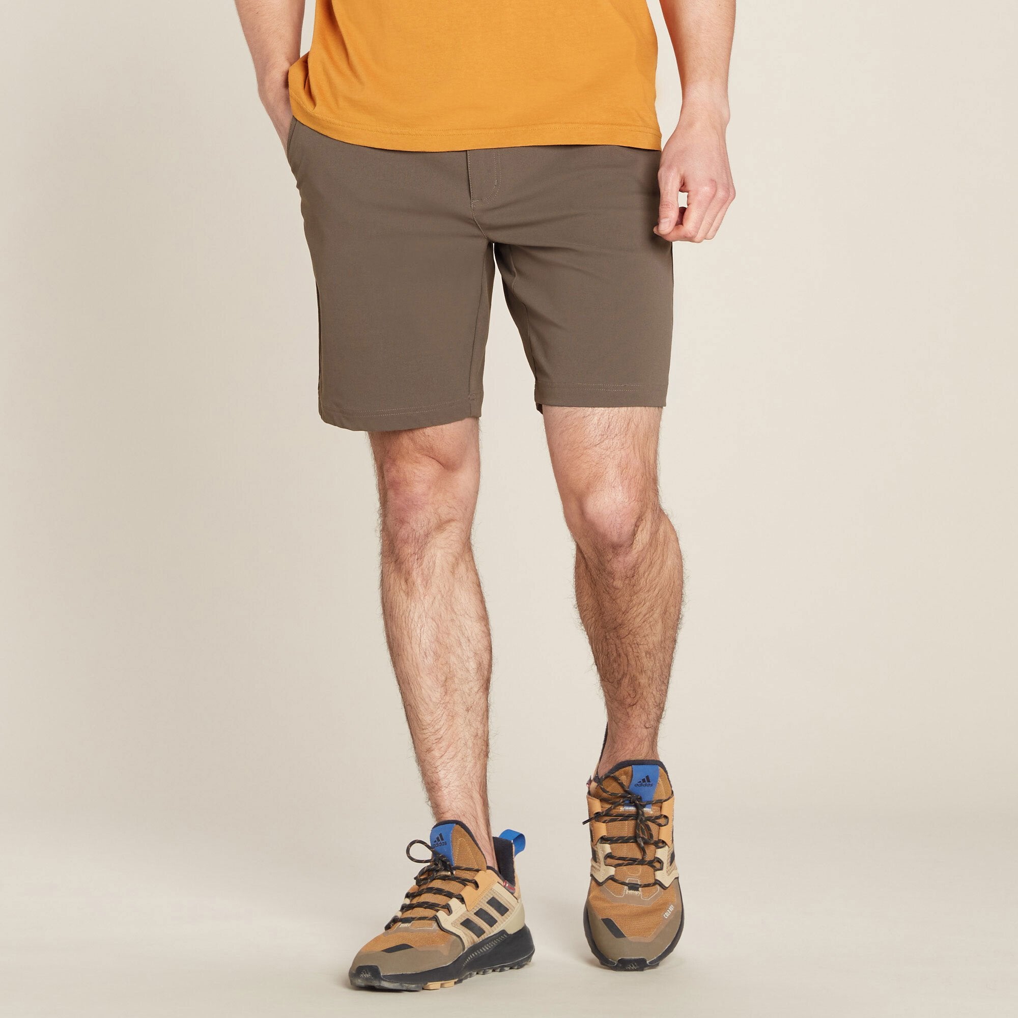 A male model wearing Sherpa Adventure Gear Bara Shorts in Green paired with a mustard-yellow t-shirt. The shorts have a tailored fit, reaching mid-thigh, with a flat-front design and no visible drawstring. He is also wearing beige and blue hiking shoes with thick soles. His stance is relaxed, with legs slightly apart and hands resting naturally by his sides. The background is a neutral beige.