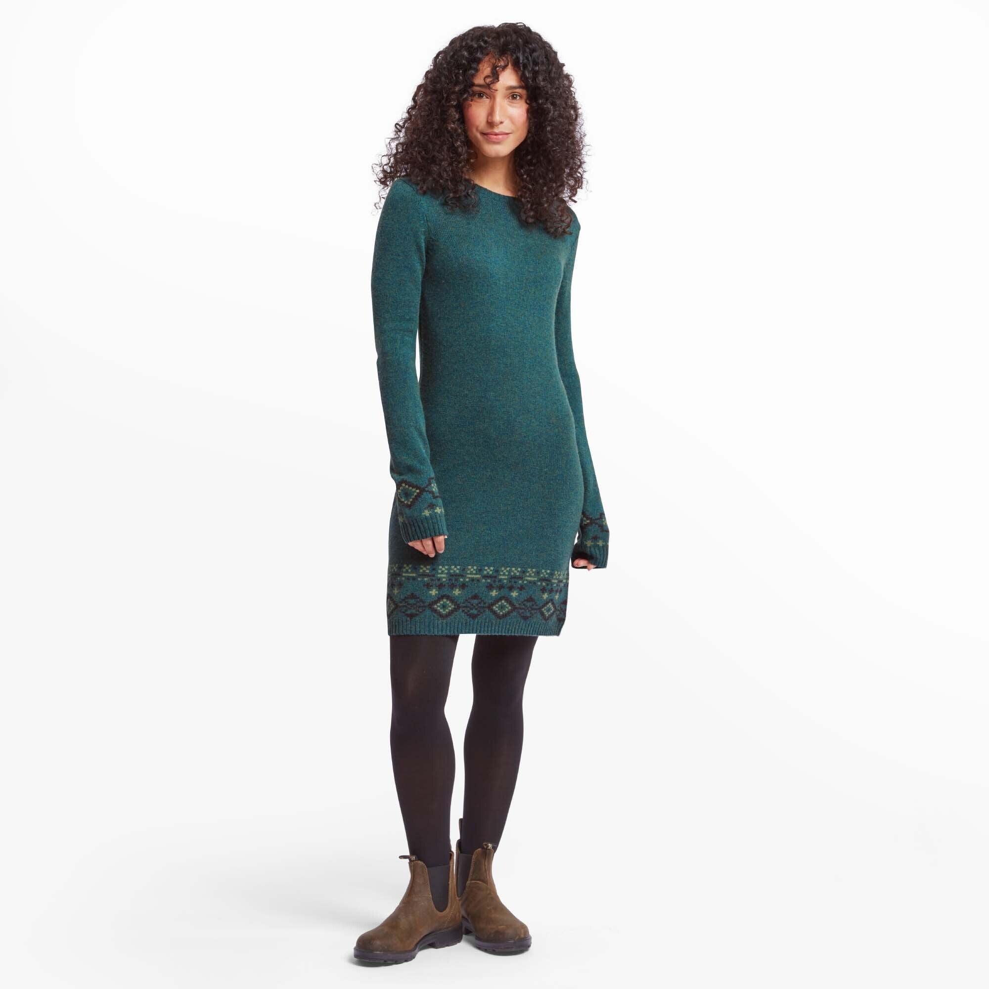 Full-length front view of a woman with curly hair wearing the Sherpa Adventure Gear Bharati Eco Dress in Green. The dress is a long-sleeve knit sweater dress with subtle heathered texture and intricate geometric patterns in dark green and lighter green at the cuffs and hemline. She pairs the dress with black tights and brown ankle boots, standing against a plain white background.