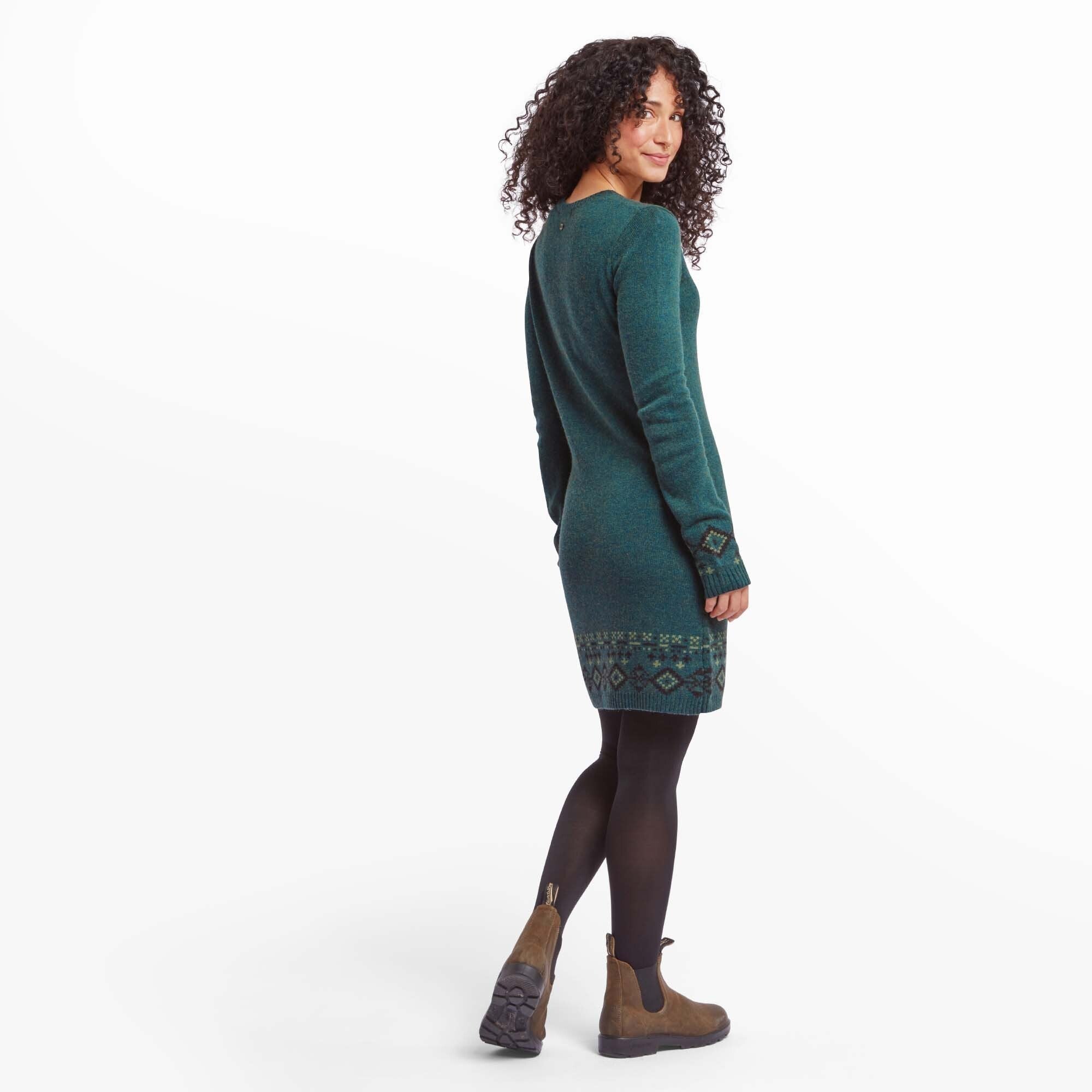 Side-angle view of the woman wearing the Sherpa Adventure Gear Bharati Eco Dress in Green. She looks back slightly, showing the dress’s comfortable fit and soft knit fabric. The side profile highlights the dress’s knee-length cut and patterned cuffs, paired with black tights and brown boots.