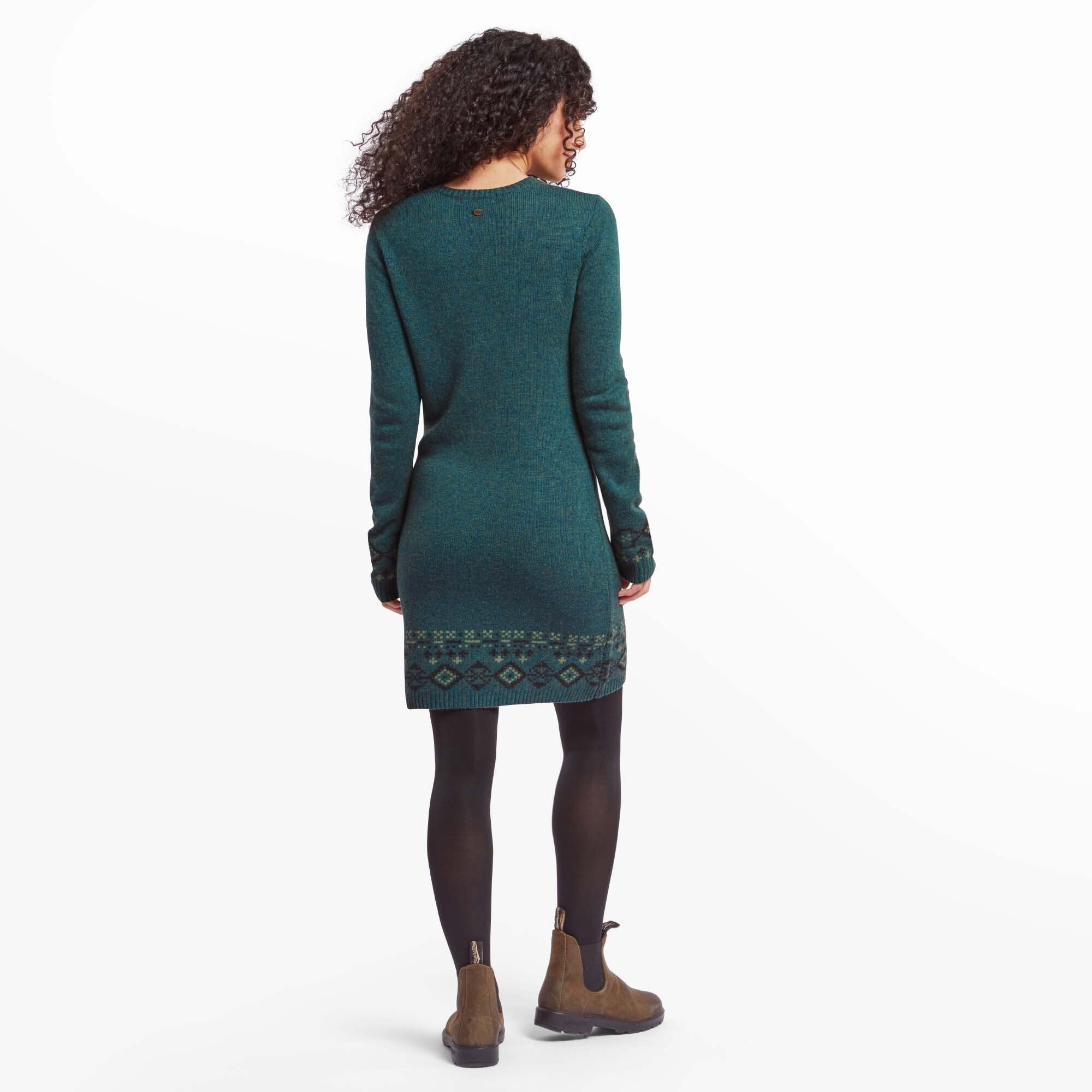 Back view of the woman modeling the Sherpa Adventure Gear Bharati Eco Dress in Green. The dress fits gently around her figure, with the patterned hemline and cuffs adding a decorative touch. Her hands rest at her sides, and her curly hair cascades down her back.