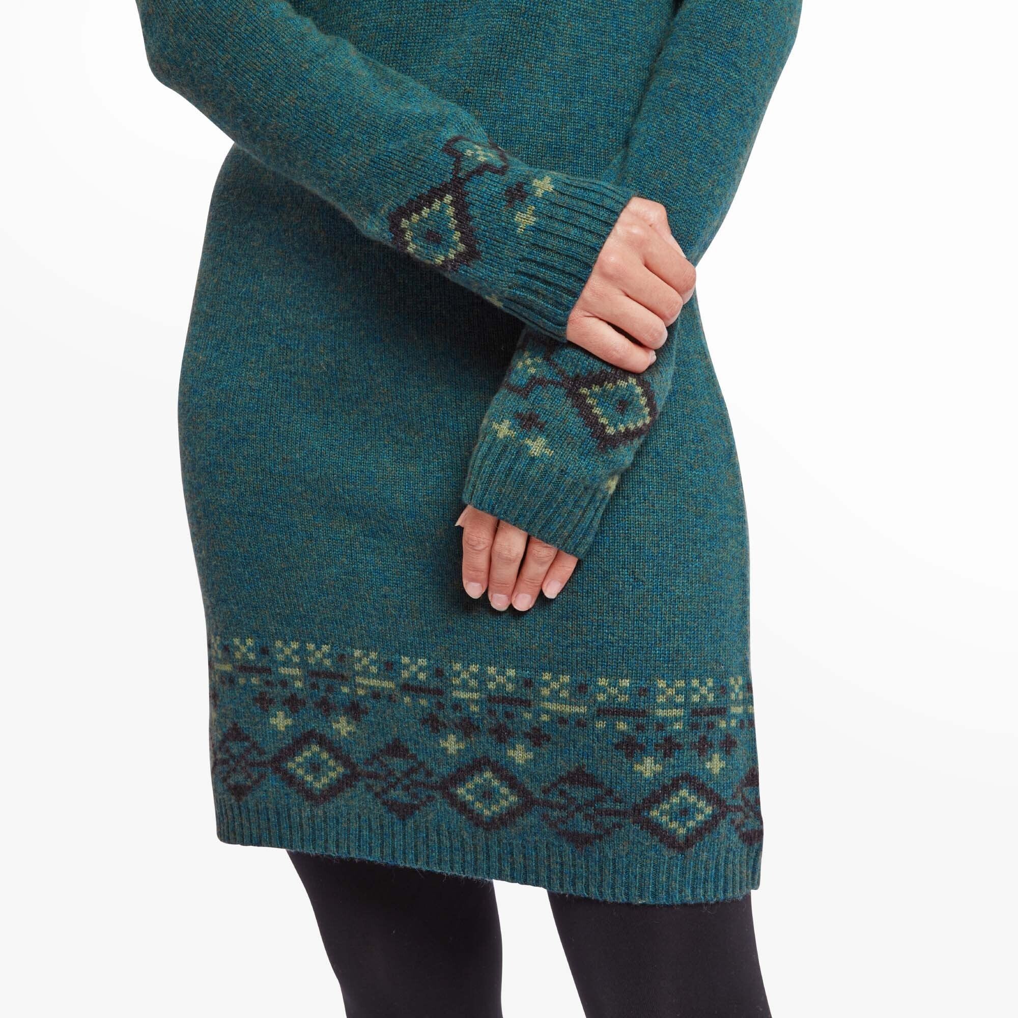 Close-up of the Sherpa Adventure Gear Bharati Eco Dress in Green’s sleeve and hemline pattern. The intricate design features geometric shapes and traditional motifs in contrasting dark green and light green hues. The soft knit texture of the fabric is visible, and her hands gently hold the edge of the sleeve, showcasing the thumbhole detail.