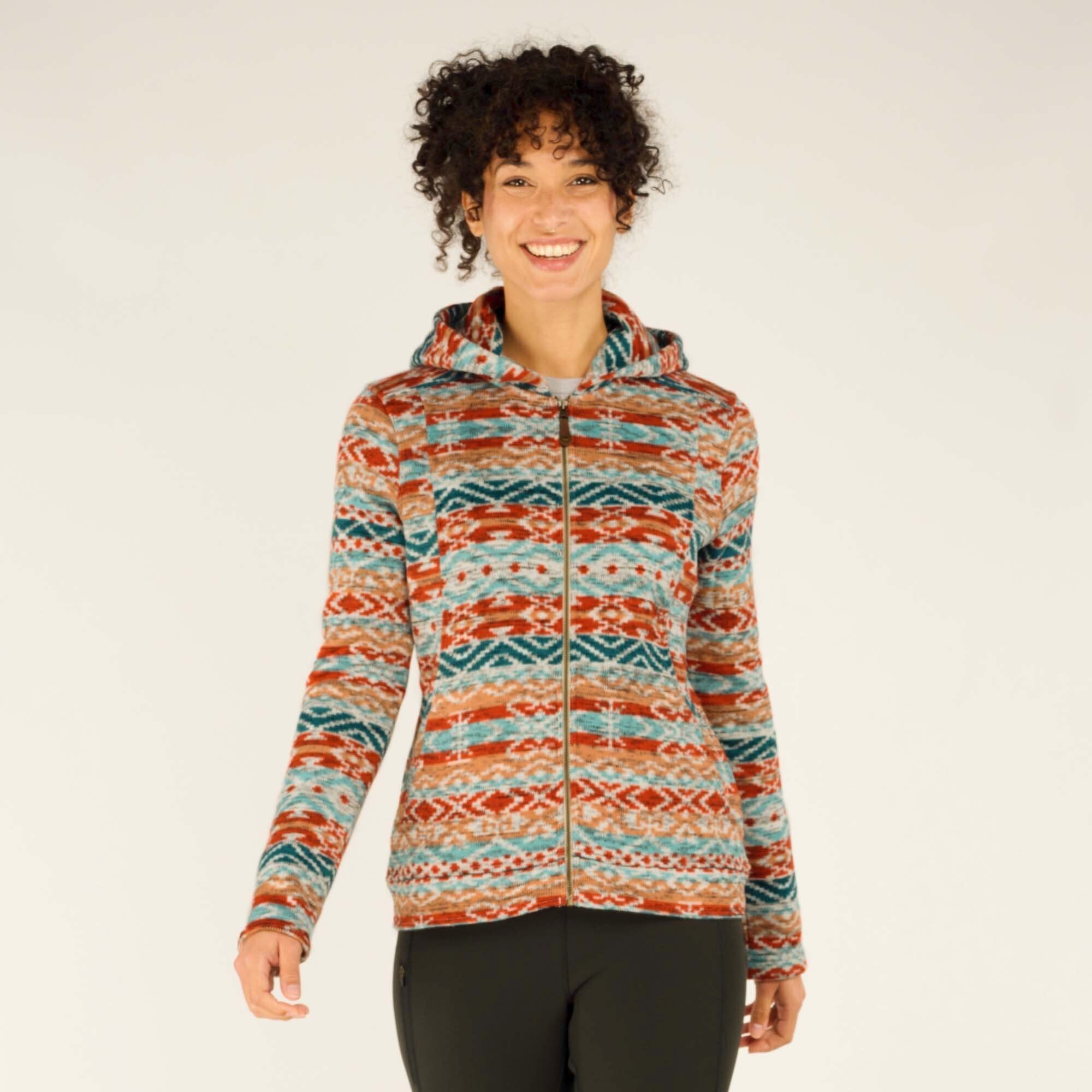 A smiling woman wearing a vibrant Sherpa Adventure Gear Bhutan Full Zip Hoodie in Beige with a colorful Bhutanese-inspired pattern of red, turquoise, and orange tones, paired with dark leggings. The hoodie features a fitted cut and a front zipper, providing a stylish and cozy appearance.