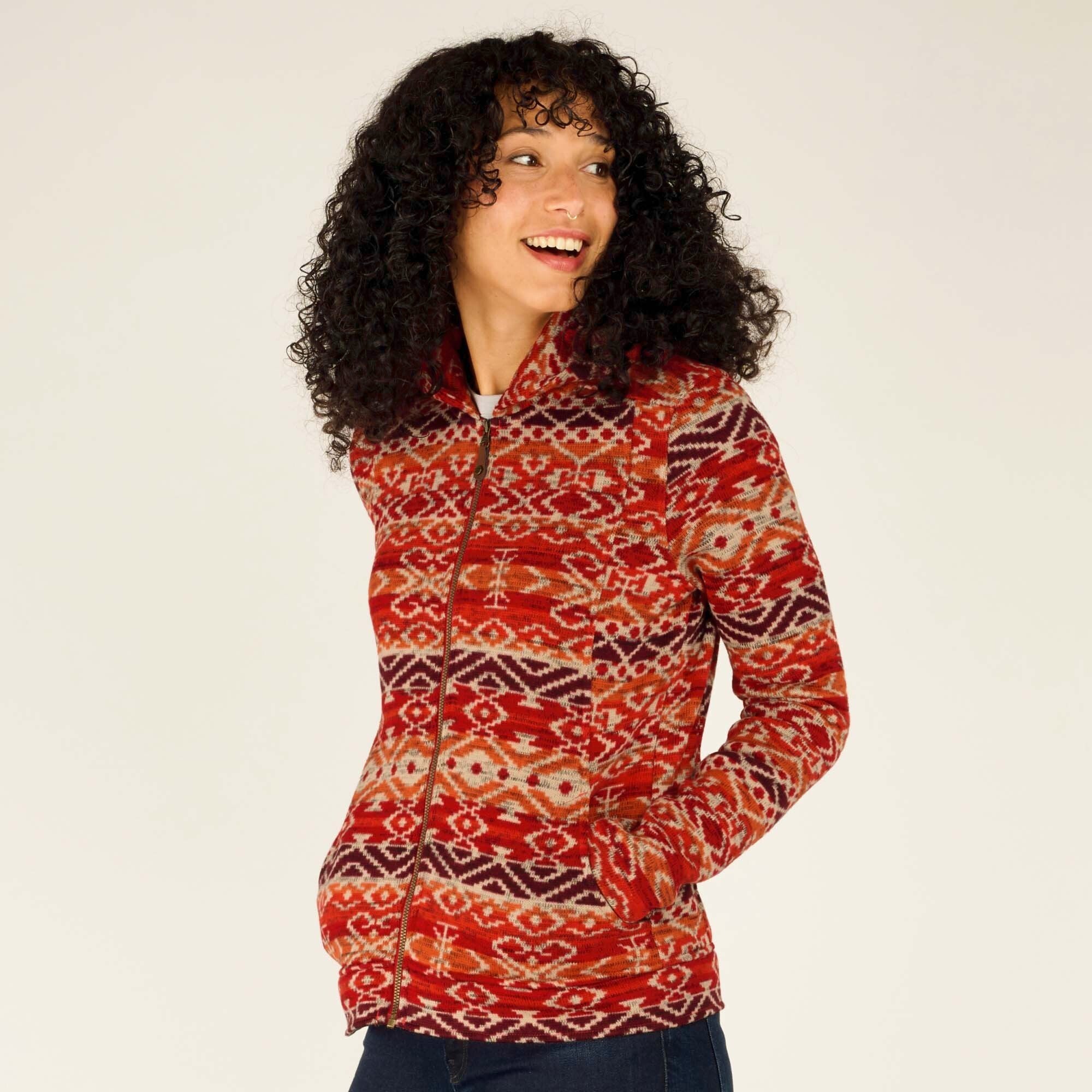 The model smiles and slightly tilts her head as she displays the Sherpa Adventure Gear Bhutan Full Zip Hoodie in Red’s front design. The intricate patterns pop against the warm tones, highlighting the craftsmanship. Her hands are casually tucked into the hoodie’s front pockets, demonstrating functionality.