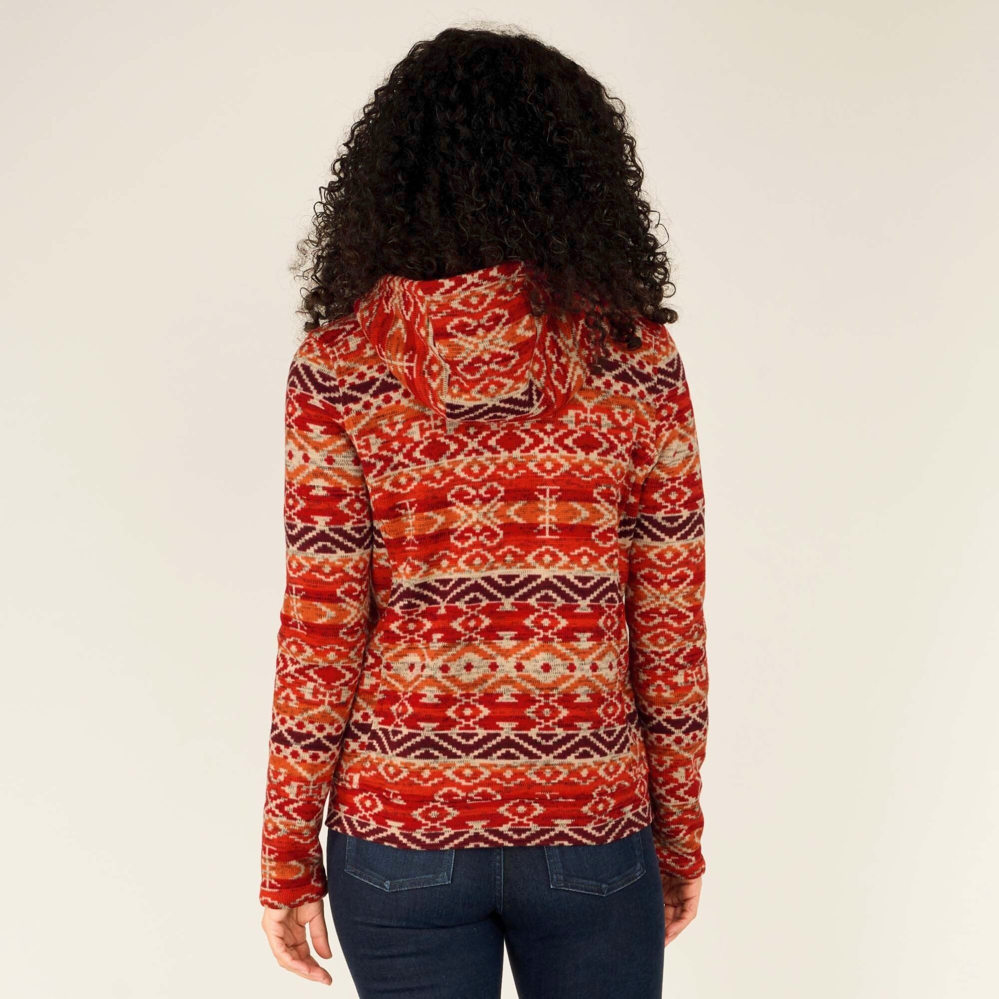 The back of the Sherpa Adventure Gear Bhutan Full Zip Hoodie in Red is fully visible, showcasing the symmetrical alignment of the Bhutanese patterns across the hood and back. The hood’s shape is clearly outlined, and the fabric’s texture suggests softness and comfort. The jeans and trainers complement the hoodie for a cohesive outfit.