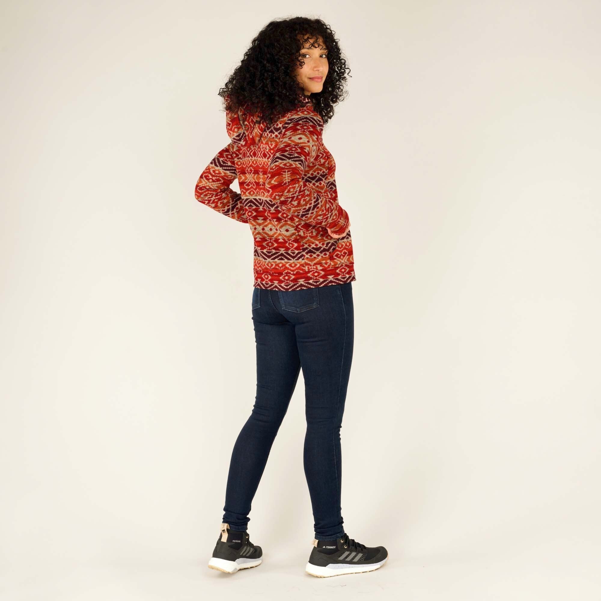 The model glances back over her shoulder with a slight smile, emphasising the Sherpa Adventure Gear Bhutan Full Zip Hoodie in Red’s versatility and stylish appeal. The side and back patterns are highlighted, while the model’s slim-fit jeans and trainers complete the casual outdoor look.