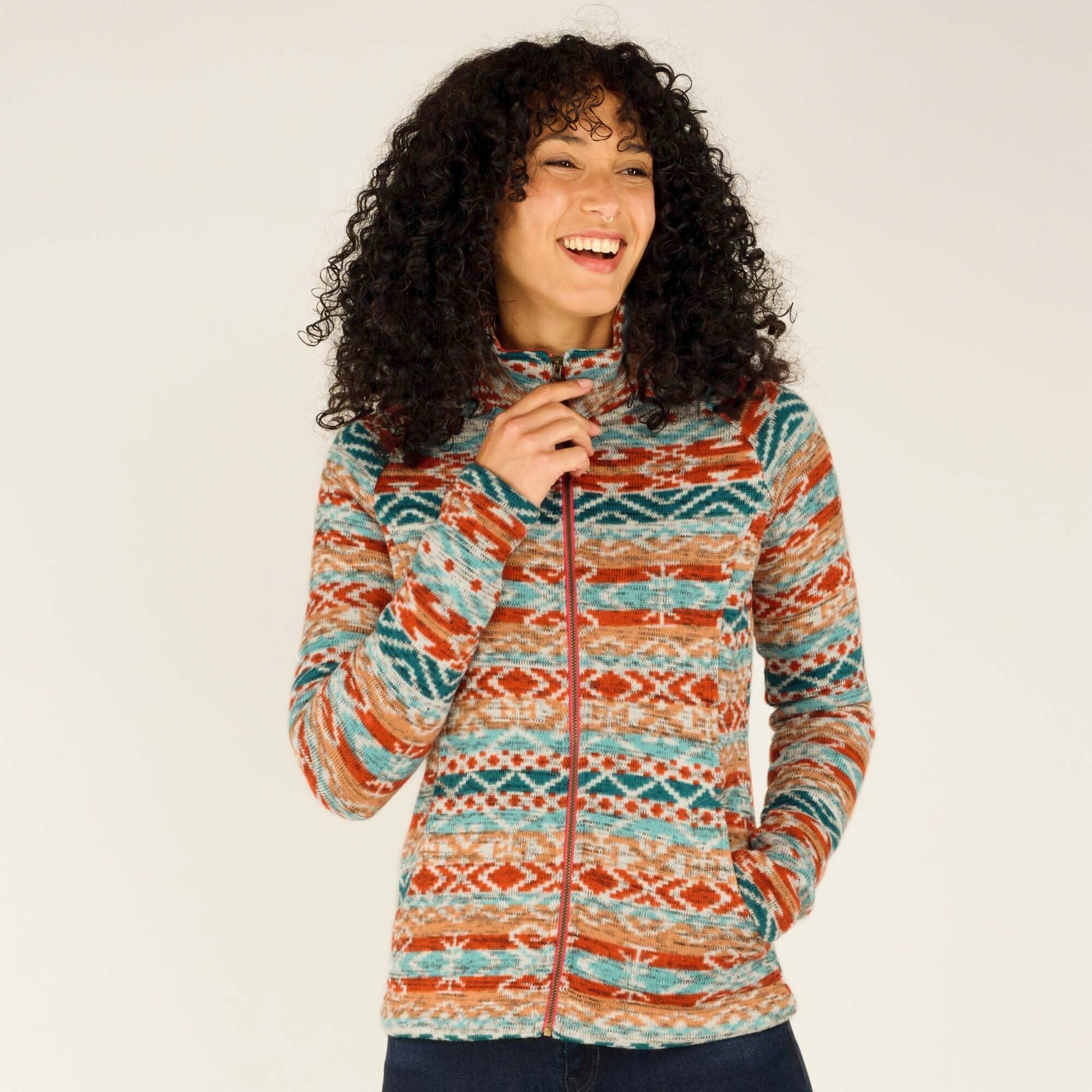 A woman wearing a Sherpa Adventure Gear Bhutan Full Zip Jacket in Beige with intricate orange, teal, and beige patterns. She smiles brightly, showcasing the high collar of the jacket, with her hand gently touching the zipper. Her curly hair frames her face as she stands against a neutral background.