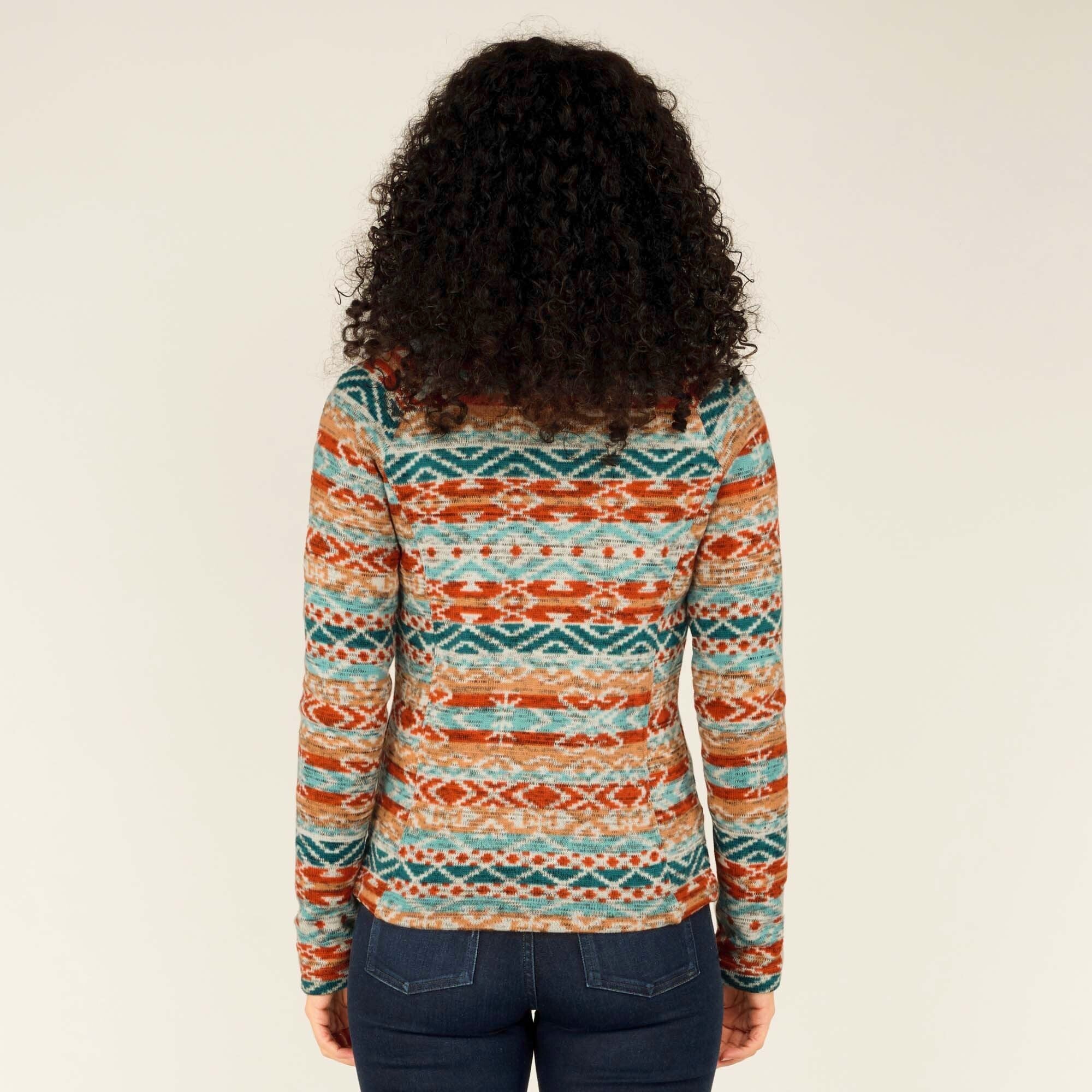 The back of the Sherpa Adventure Gear Bhutan Full Zip Jacket in Beige is displayed, showcasing the all-over colorful pattern of geometric shapes and lines in orange, teal, and beige hues. Her curly hair cascades down her back, partially covering the collar.