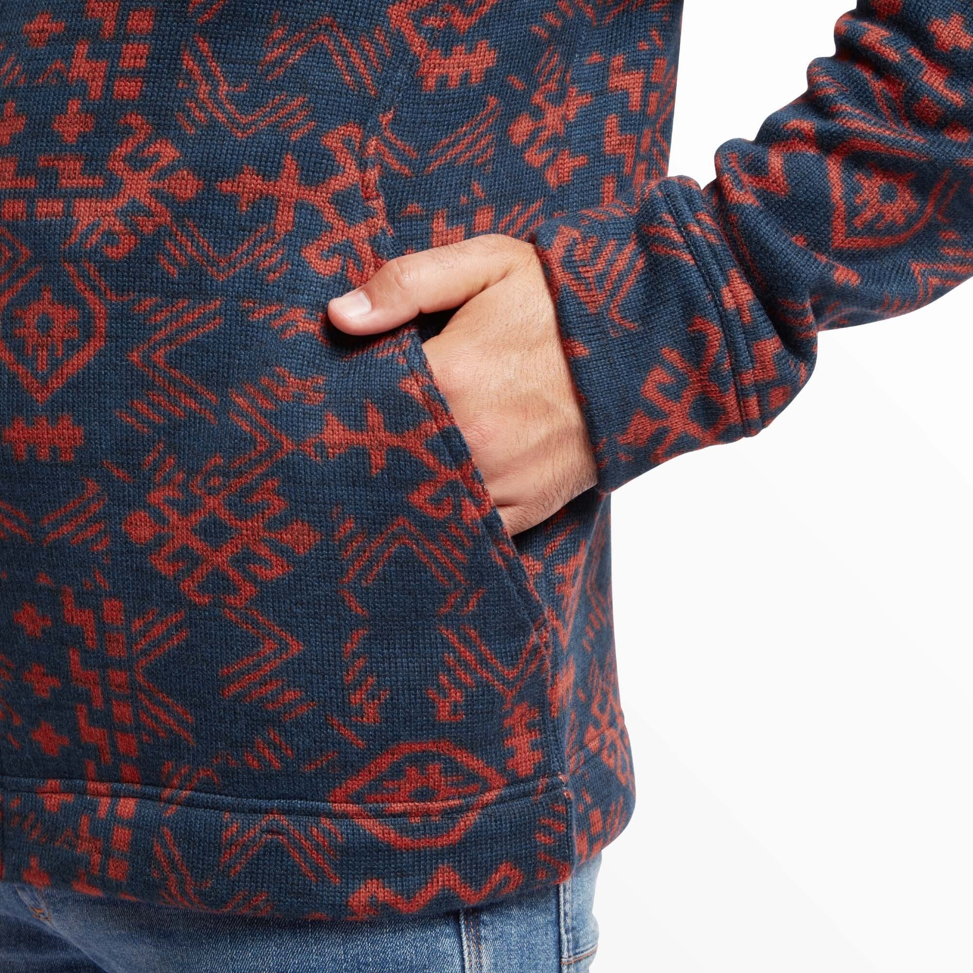 Close-up of a hand placed in the pocket of a Sherpa Adventure Gear Bhutan Full Zip Jacket in Blue with a vibrant red geometric pattern. The fabric texture is slightly ribbed, emphasising the woven detail of the design. The pocket is seamlessly integrated into the pattern, and the sleeve hem appears snug, highlighting the craftsmanship. The bottom edge of the jacket is visible, resting just above a pair of light blue denim jeans, which add a casual touch to the look.