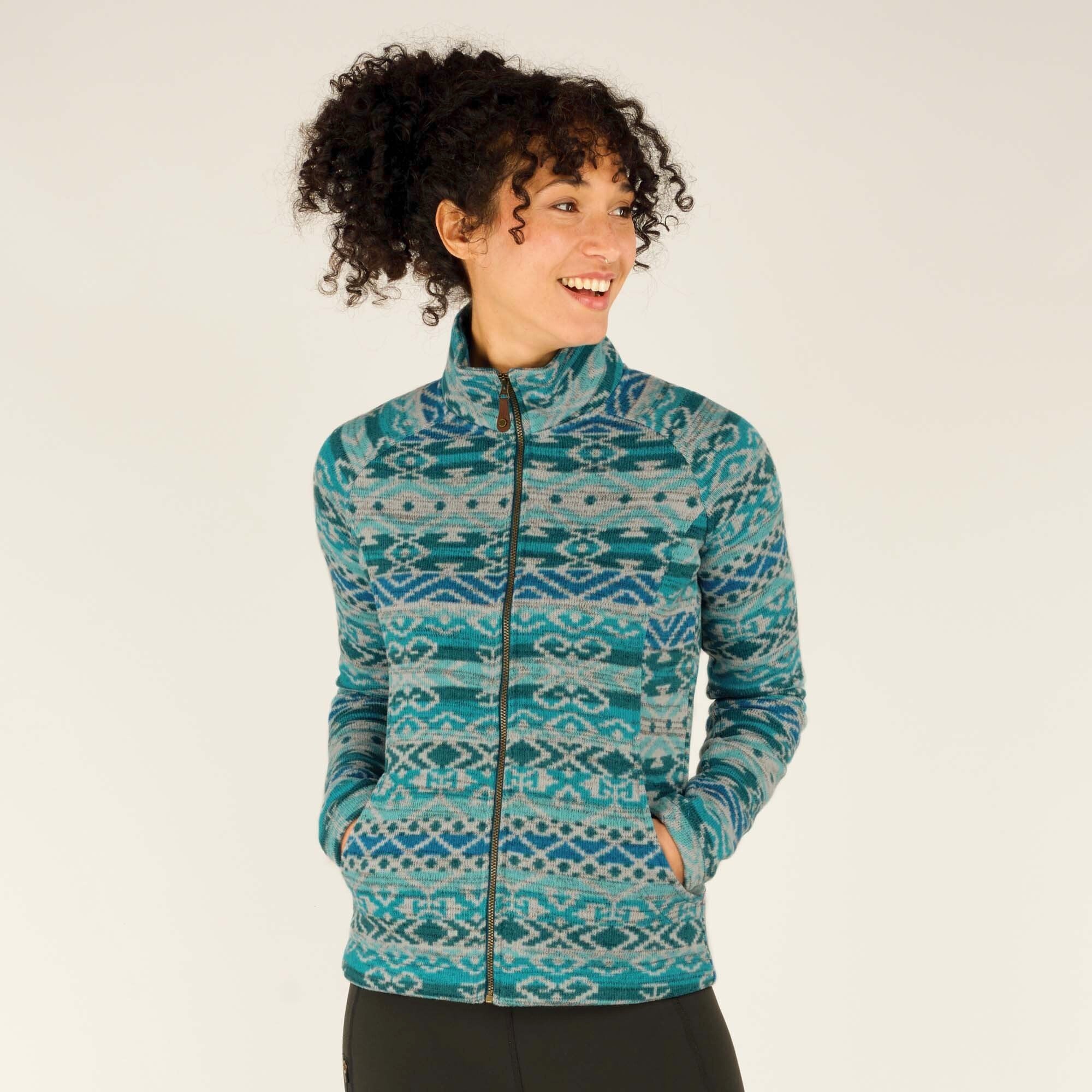 A woman with curly dark brown hair smiles while wearing a Sherpa Adventure Gear Bhutan Full Zip Jacket in Green. The jacket features intricate geometric designs in shades of blue and teal, a high collar, and a front zipper. She has her hands tucked into the side pockets and is wearing black leggings. The background is a neutral off-white.