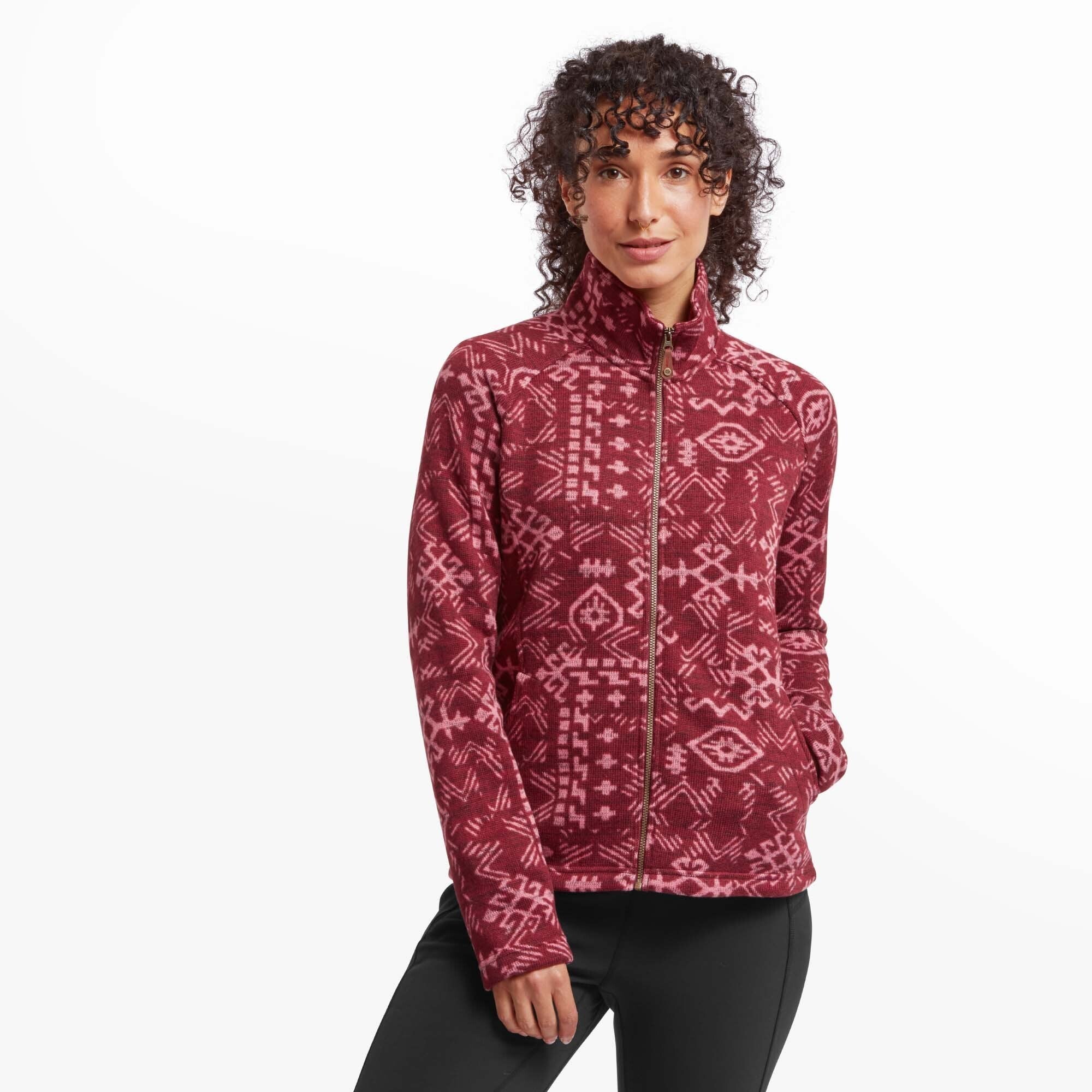 A female model with curly hair wears the Sherpa Adventure Gear Bhutan Full Zip Jacket in Red with an intricate Himalayan-inspired geometric pattern in a lighter pink hue. She stands with her hands in her jacket pockets, looking directly at the camera with a confident and relaxed expression. The jacket features a full front zipper, a high collar, and a cozy fleece texture. She pairs the jacket with black leggings.