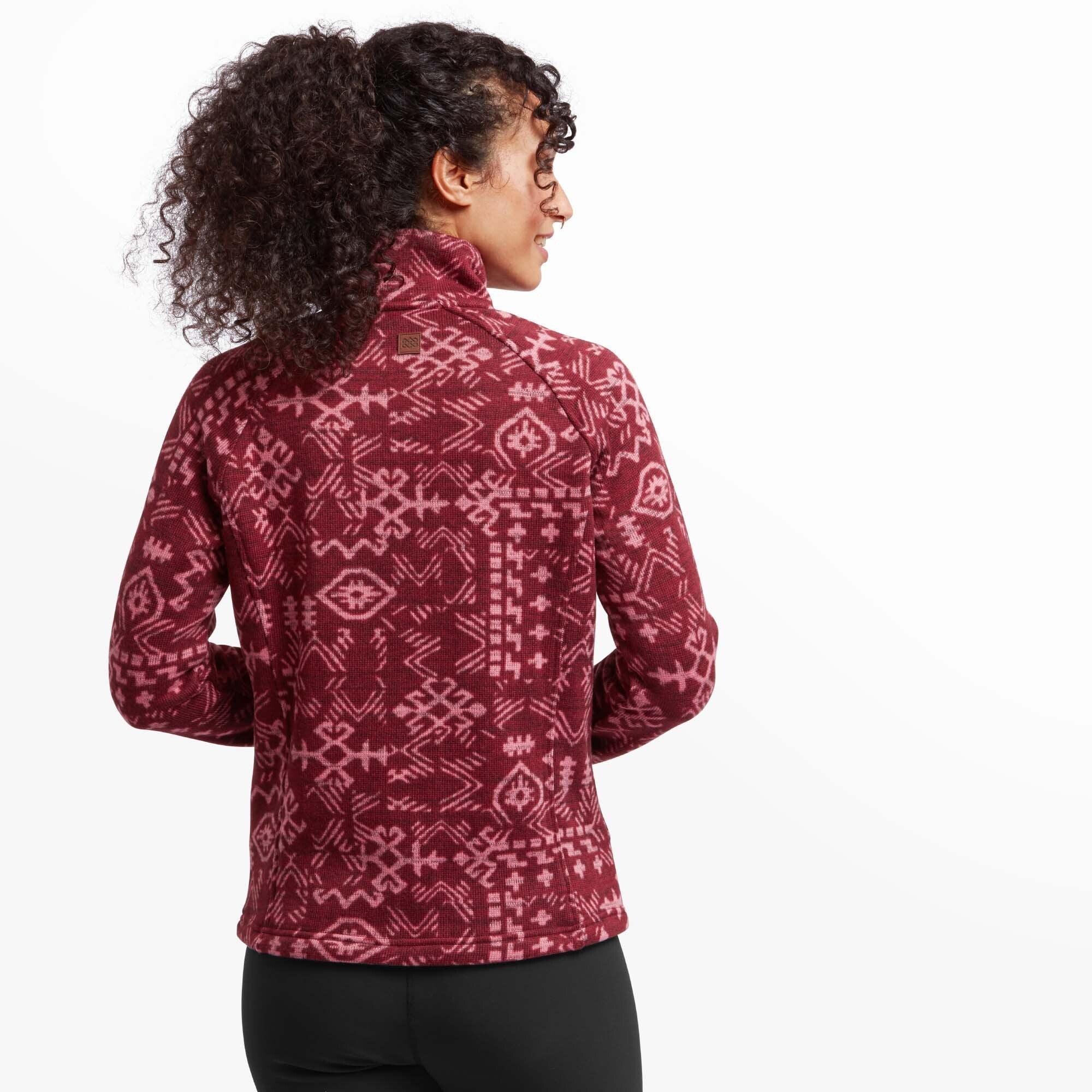 A back view of the model wearing the Sherpa Adventure Gear Bhutan Full Zip Jacket in Red, showing the rear design. The red jacket is covered in the same geometric Himalayan-inspired pattern, and the high collar provides added warmth. A small Sherpa logo patch is visible at the upper back, just below the collar.