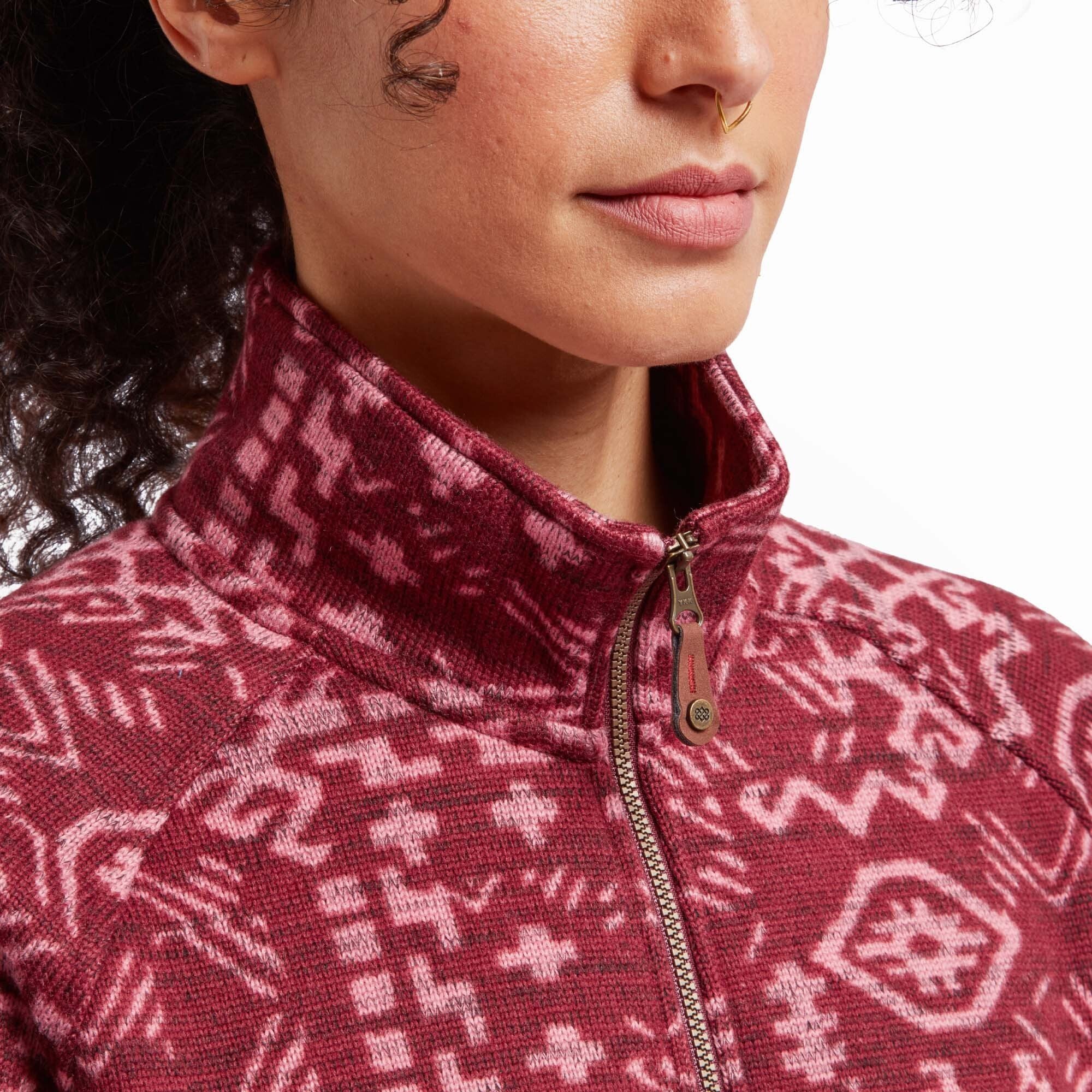 A close-up of the Sherpa Adventure Gear Bhutan Full Zip Jacket in Red’s collar and zipper. The high collar is fully zipped, and the woven pattern continues seamlessly into the collar. The zipper pull features a small Sherpa logo charm, adding a branded detail. The fleece texture of the fabric is clearly visible, showing the soft and warm quality of the material.