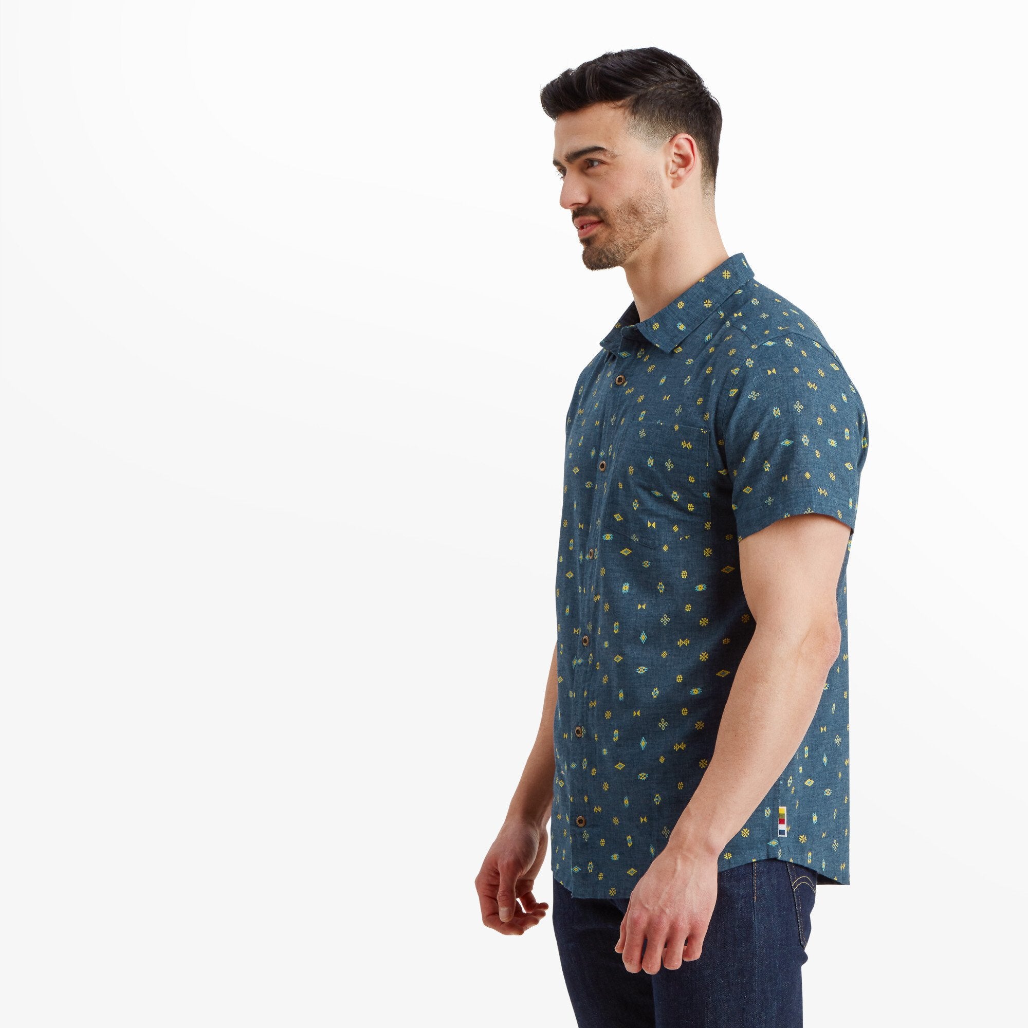 The same man is shown from a slight side angle of the Sherpa Adventure Gear Bhutan Geo Short Sleeve Shirt in Blue, highlighting the fit of the shirt on his upper torso and shoulders. The short sleeves and hemline are clearly visible, providing a relaxed and comfortable look. The small geometric designs maintain their consistent pattern across the shirt.