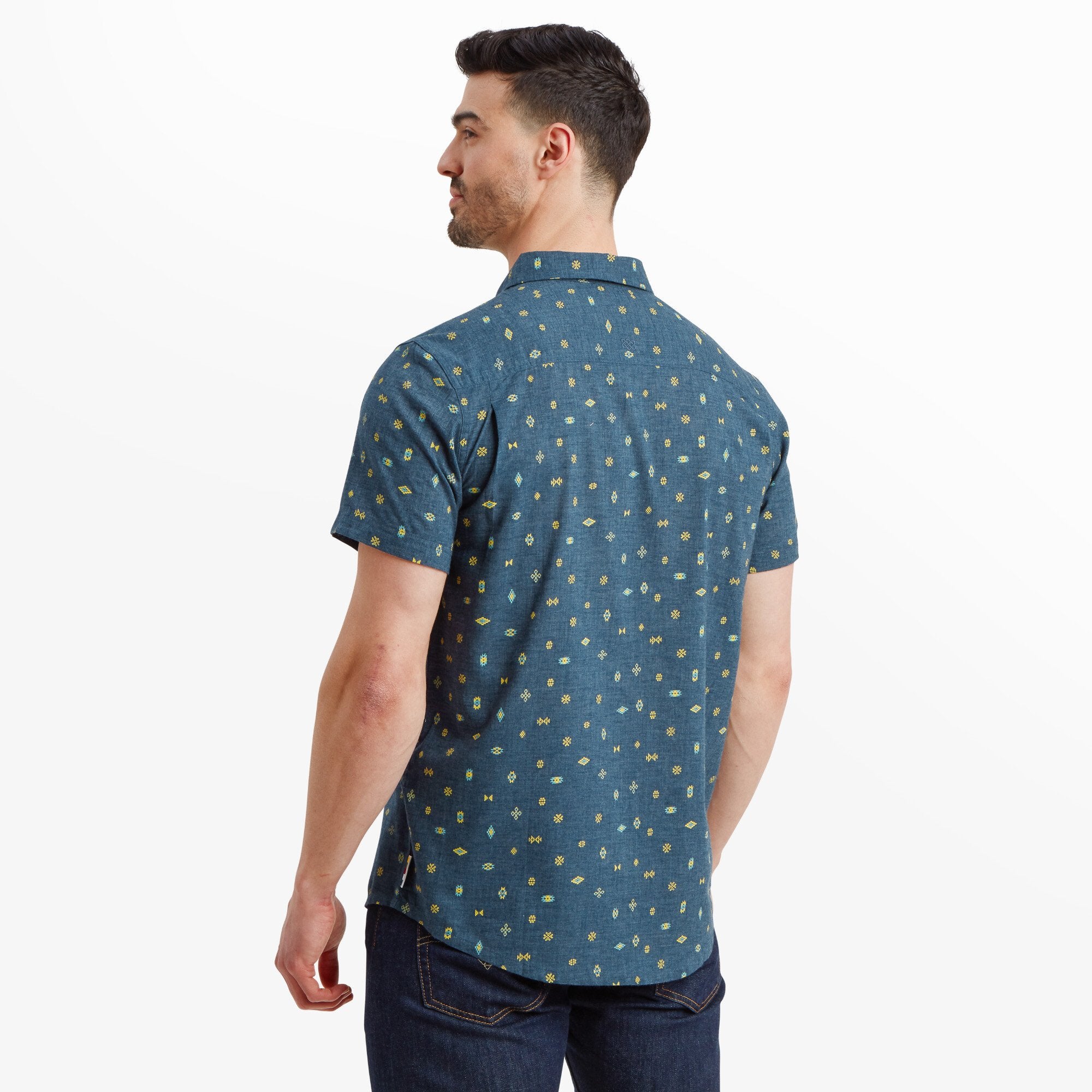 A rear view of the man shows the back of the Sherpa Adventure Gear Bhutan Geo Short Sleeve Shirt in Blue, continuing the geometric pattern seamlessly. The curved hemline adds to the casual and versatile style of the shirt. His posture highlights the shirt's tailored yet relaxed fit.